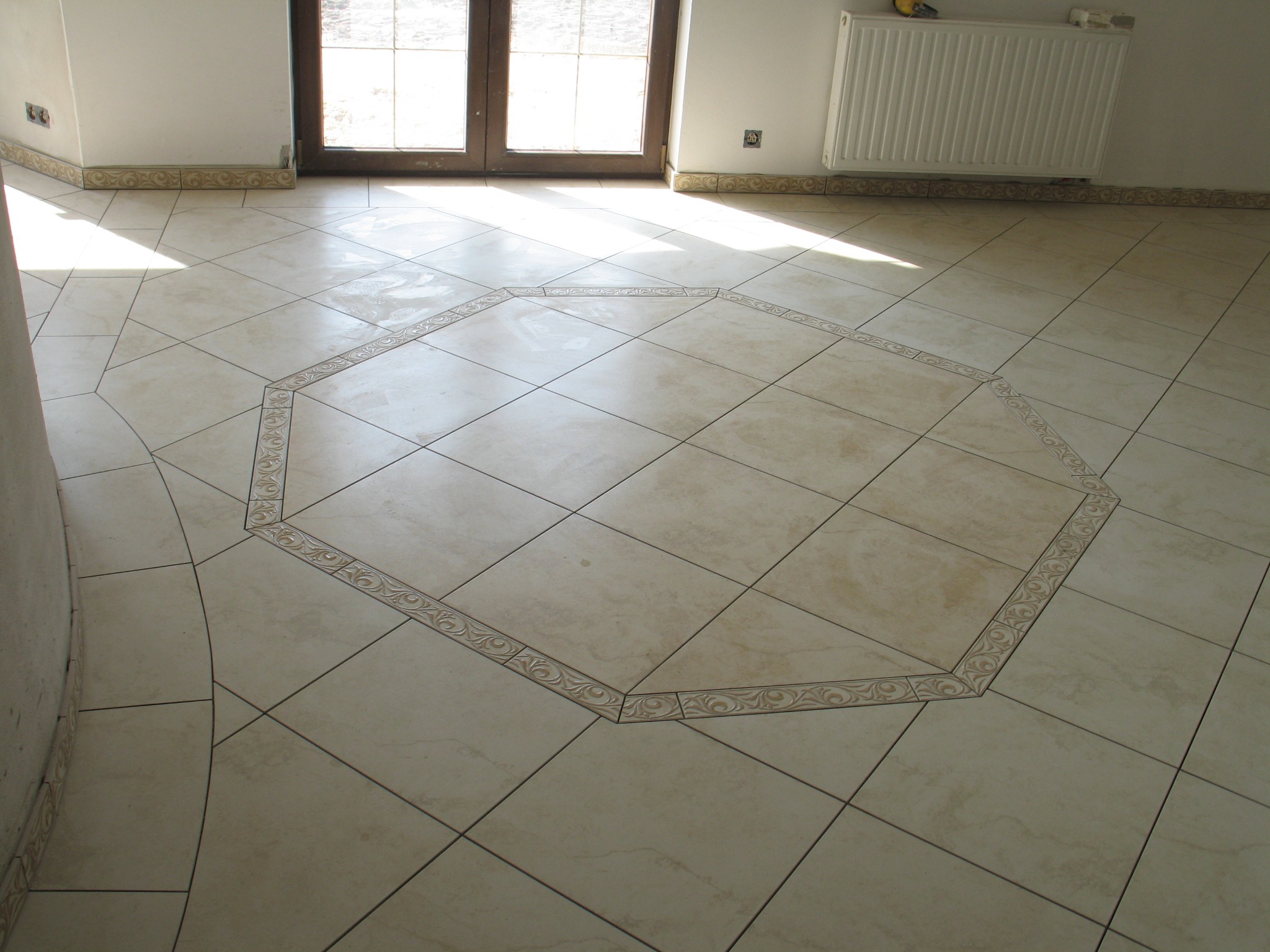 Beżowa podłoga z płytek ceramicznych ułożonych w kwadraty i oktagonalny wzór z dekoracyjnym obramowaniem, jasne wnętrze z oknem balkonowym i grzejnikiem.