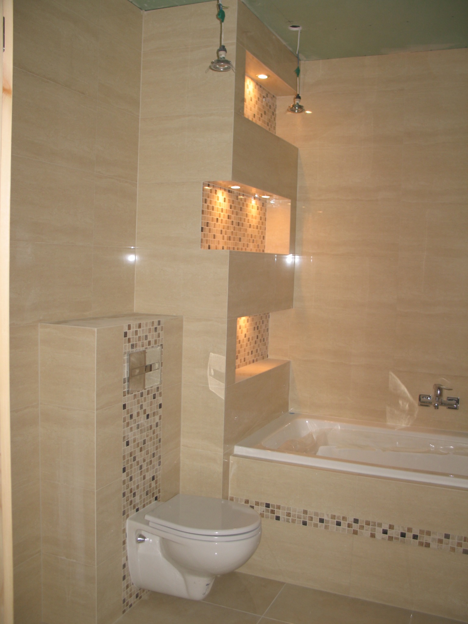 Wykończona łazienka z beżowymi płytkami, dekoracyjnymi mozaikami, podświetlanymi niszami i podwieszaną toaletą.