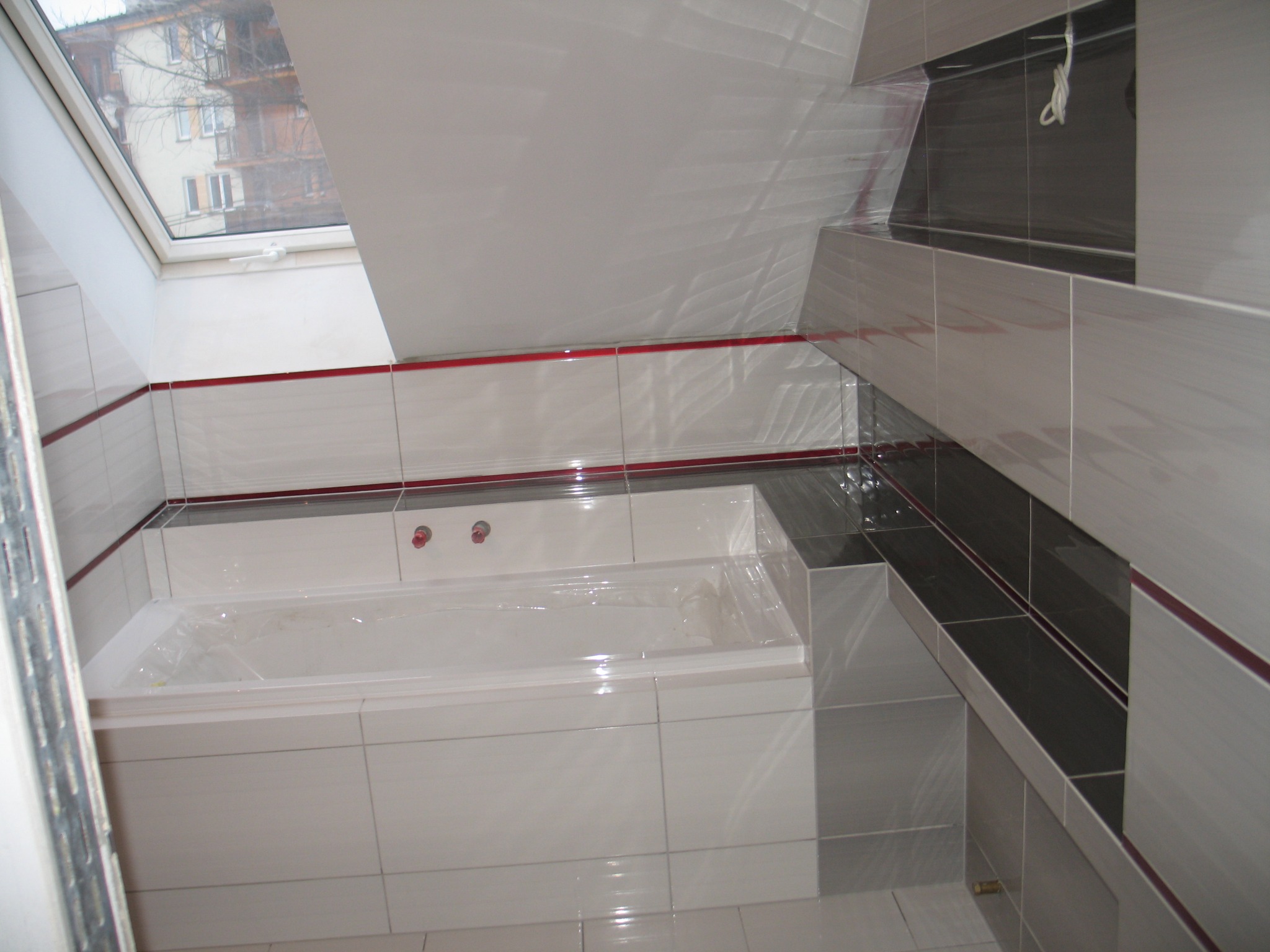 Wykończona łazienka na poddaszu z biało-szarymi płytkami, czerwoną fugą i oknem dachowym, widoczna wanna zabezpieczona folią ochronną.