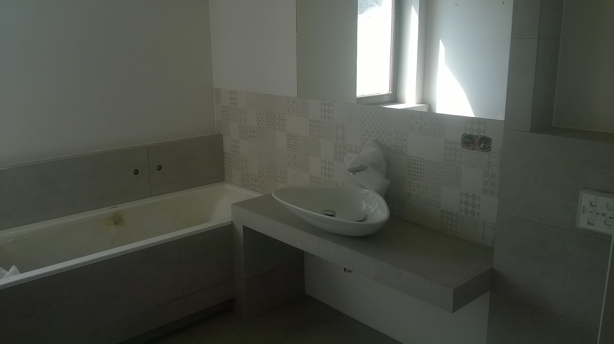 Nowoczesna łazienka z wanną i umywalką nablatową; płytki ścienne w geometryczne wzory, szare panele obudowy wanny i blatu umywalki, okno z widocznym światłem dziennym, w tle surowe gniazdka...