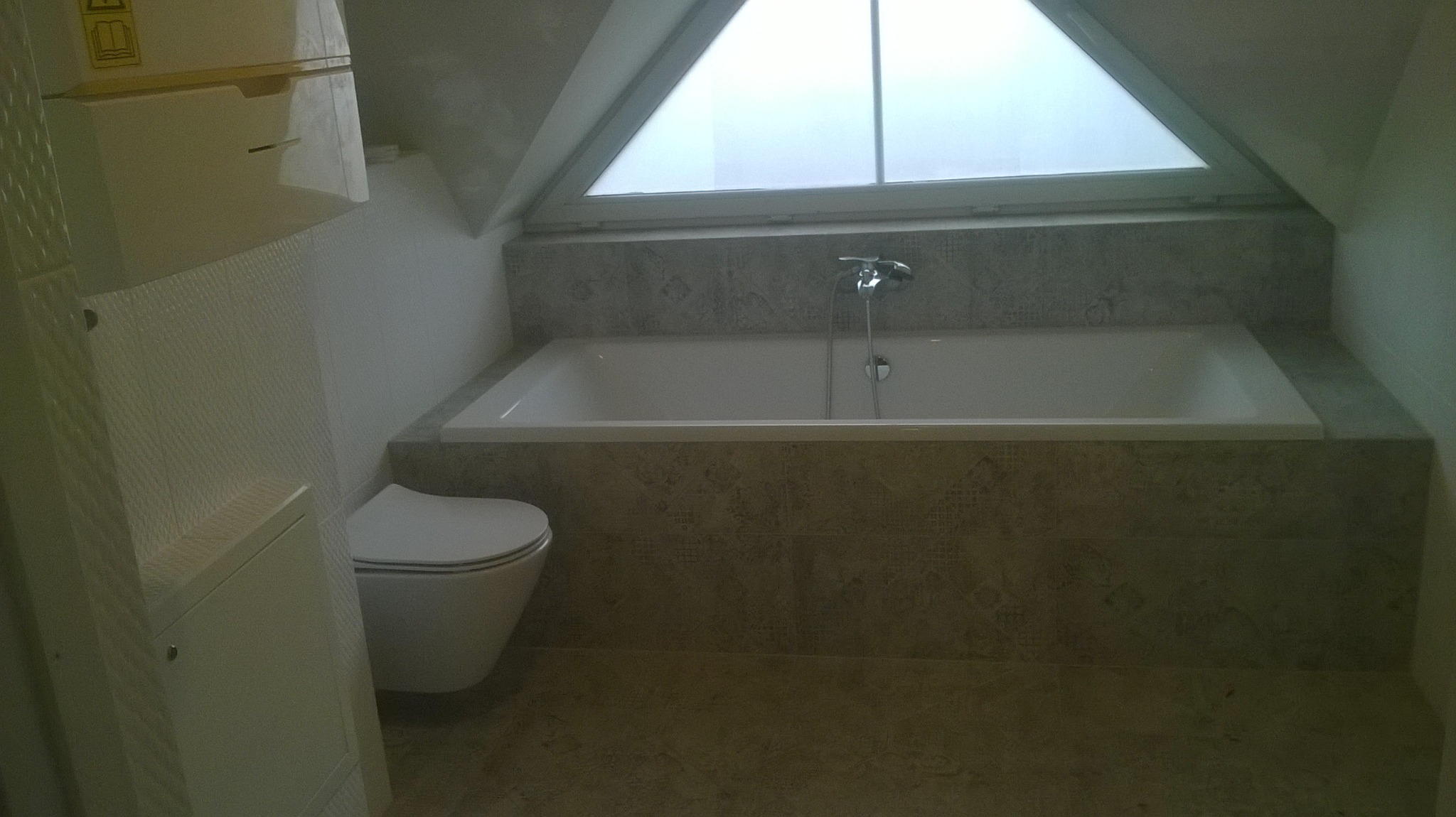 Wykończona łazienka na poddaszu z wanną obudowaną płytkami w odcieniach szarości, wiszącą toaletą i oknem dachowym w kształcie trójkąta.