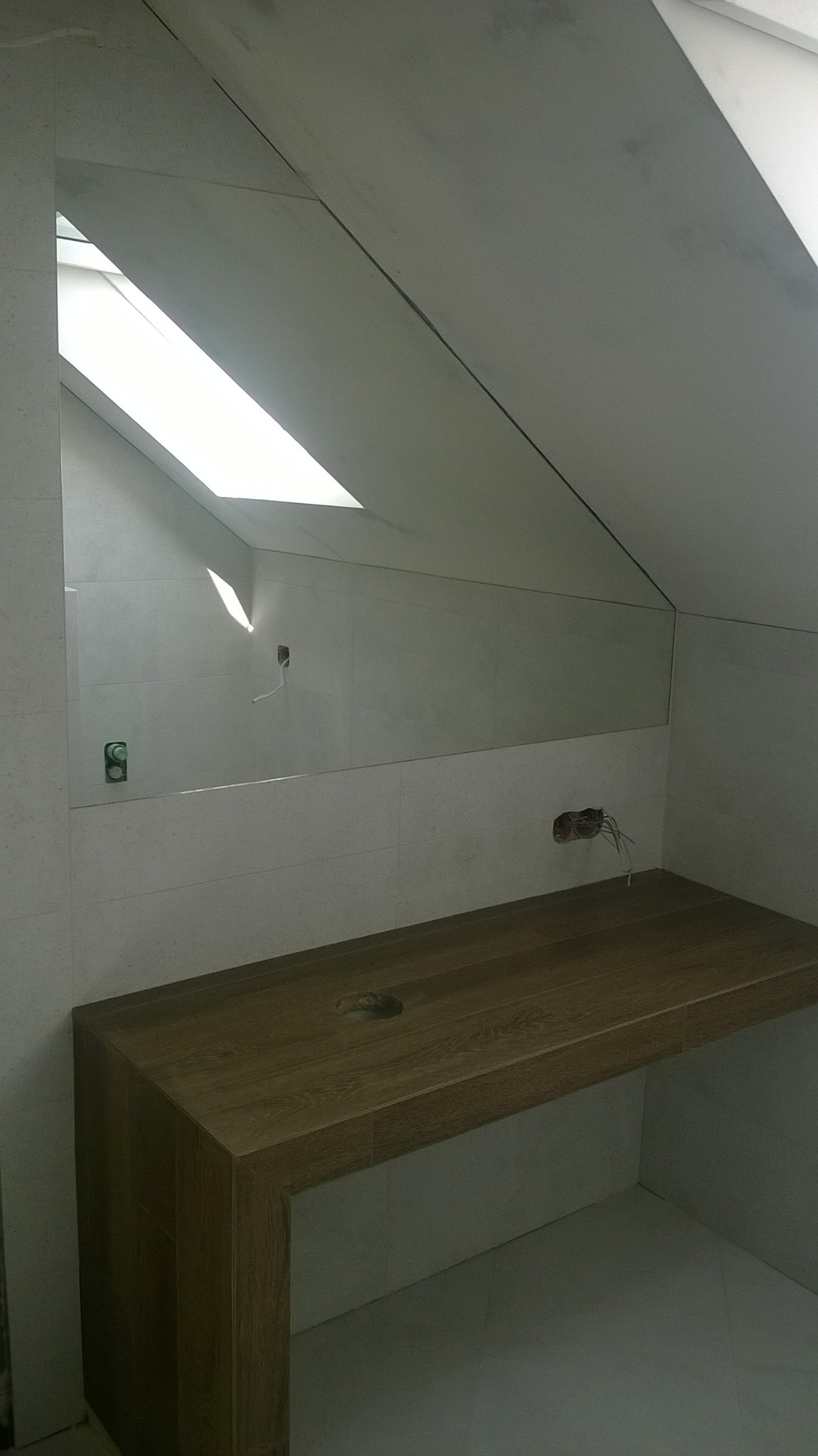 Łazienka na poddaszu w trakcie wykańczania: lustro bez ramy, drewniany blat z otworem na umywalkę, białe płytki na ścianach, widoczne gniazdka i wystające kable.