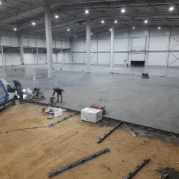 Wnętrze hali przemysłowej w trakcie wykonywania wylewki. Widoczny pracownik zacierający powierzchnię, zapas worków z materiałem, listwy dylatacyjne oraz samochód dostawczy z logo firmy.