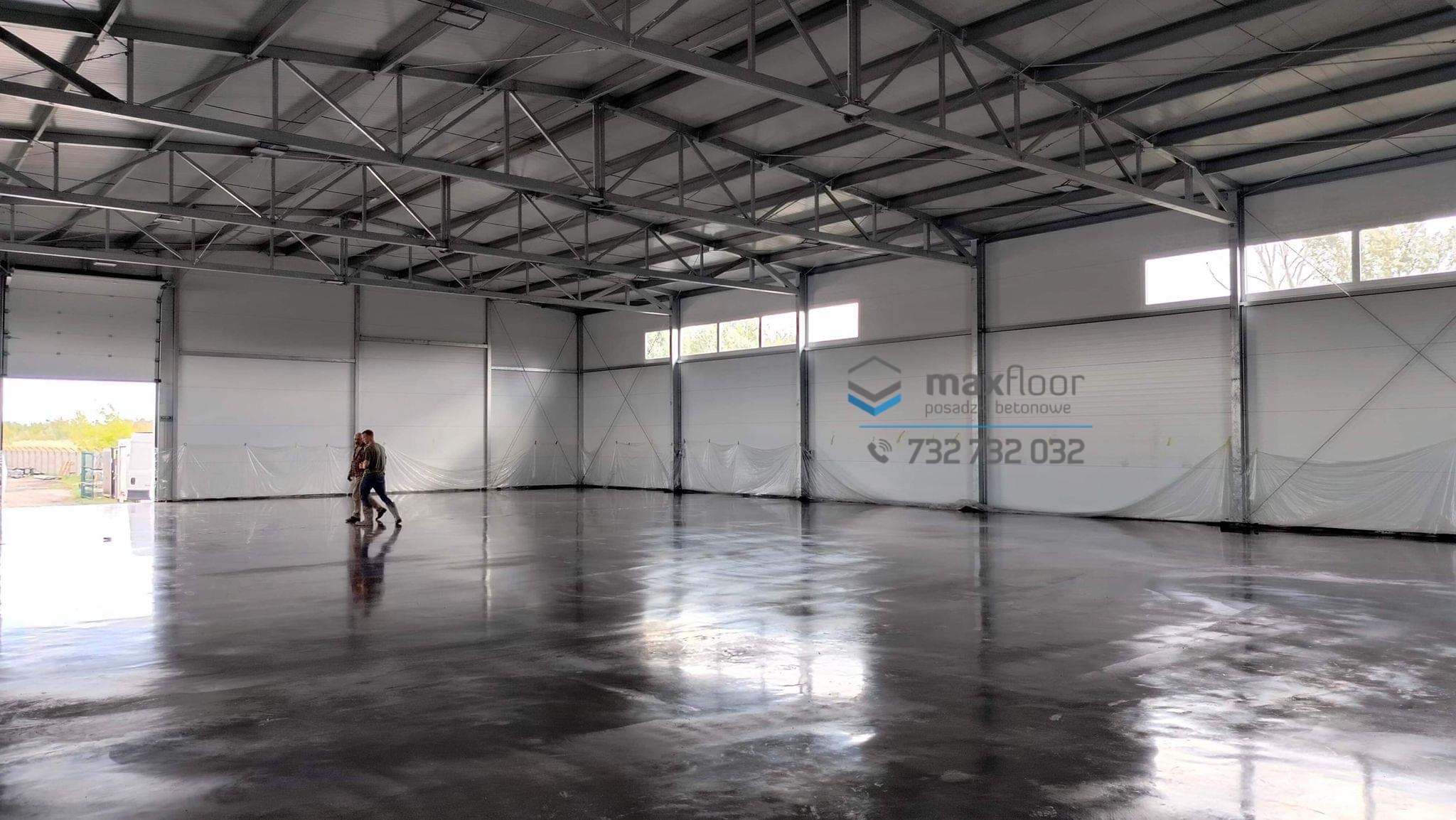 Błyszcząca, szara podłoga w hali przemysłowej z widocznymi dwoma osobami i logo firmy Maxfloor na ścianie.