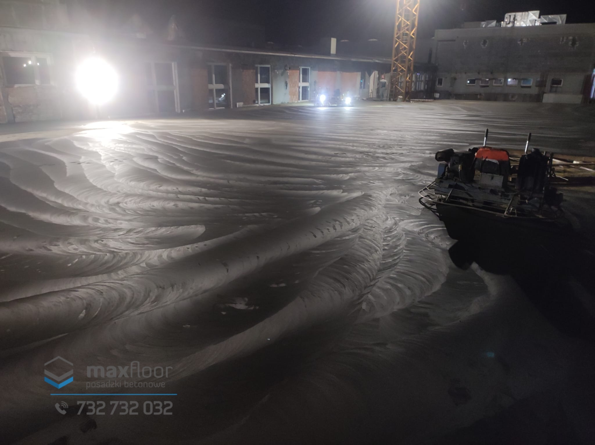 Wygładzanie świeżo wylanej betonowej posadzki przemysłowej z użyciem zacieraczki mechanicznej, widoczne ślady obróbki i elementy konstrukcyjne budynku w tle pod nocnym oświetleniem.