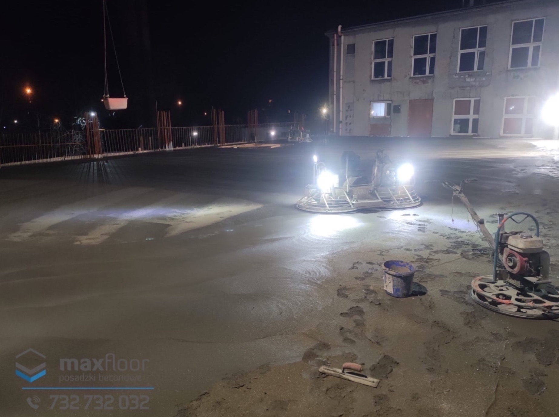 Nocna praca przy wylewaniu betonu na budowie. Dwie zacieraczki mechaniczne do betonu oświetlają plac budowy, widoczne wiadro i kielnia. W tle budynek w stanie surowym.