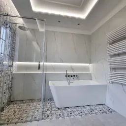 Nowoczesna łazienka z białą wanną, szklanym prysznicem i dekoracyjnymi płytkami podłogowymi w geometryczny wz&oacute;r, oświetlenie LED w suficie i wnęce ściennej.