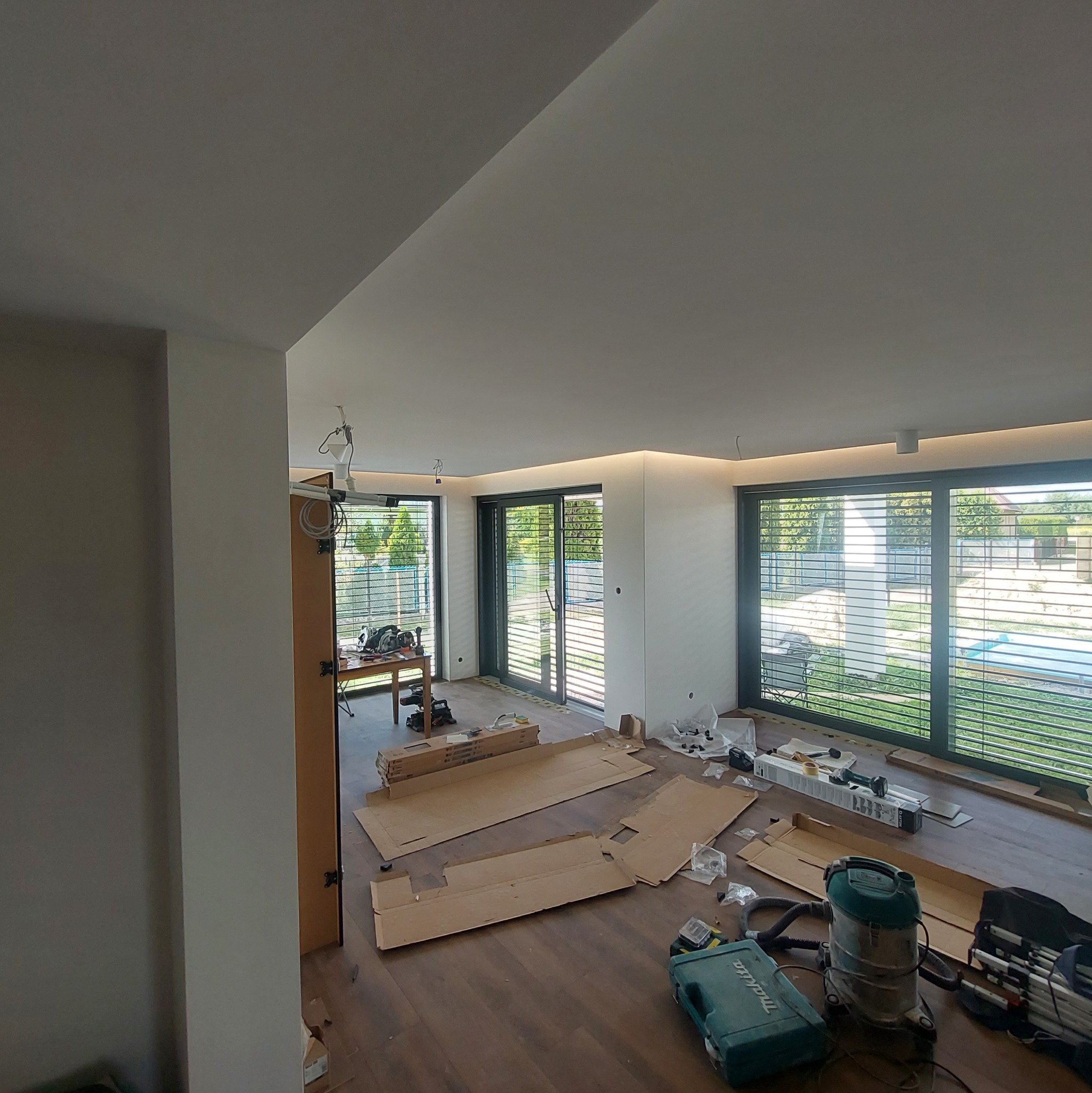 Wnętrze w trakcie wykańczania: panele podłogowe, narzędzia, kartony, duże okna z żaluzjami. Widoczne oświetlenie sufitowe LED.