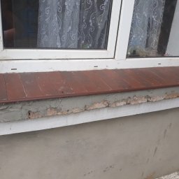 Sasza Korets - Widok z bliska na parapet zewnętrzny z brązowych płytek, szarej zaprawy i białej listwy, pod oknem PCV; fragment ściany z szarą strukturą.