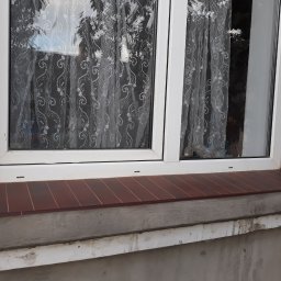 Sasza Korets - Białe okno PCV z brązowym parapetem wykończonym płytkami ceramicznymi, widoczne ślady naprawy muru pod parapetem i firanka w oknie.