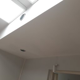 Sasza Korets - Wnętrze po remoncie, widok na gładki, pomalowany sufit z okrągłymi otworami wentylacyjnymi, górne okno dachowe i fragment ściany z drzwiami.