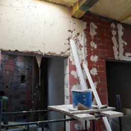 Sasza Korets - Częściowo otynkowana ściana z czerwonej cegły wewnątrz budynku w trakcie prac, widoczny fragment rusztowania, wiadro z zaprawą i narzędzia na prowizorycznym blacie, oraz fragment sylwetki...