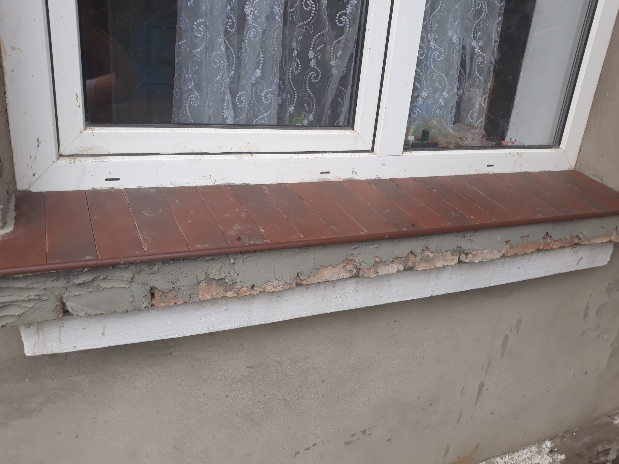 Widok z bliska na parapet zewnętrzny z brązowych płytek, szarej zaprawy i białej listwy, pod oknem PCV; fragment ściany z szarą strukturą.