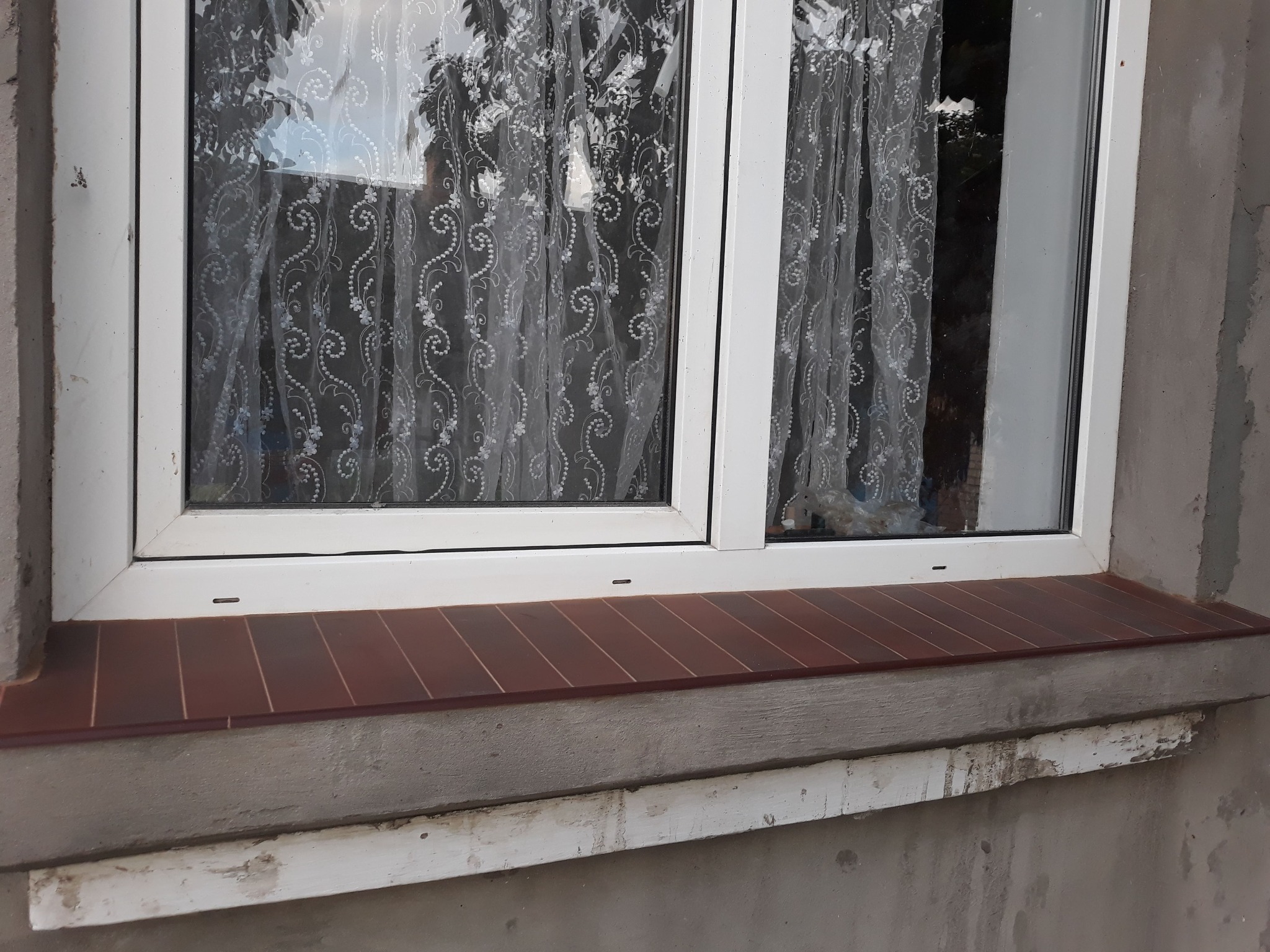 Białe okno PCV z brązowym parapetem wykończonym płytkami ceramicznymi, widoczne ślady naprawy muru pod parapetem i firanka w oknie.