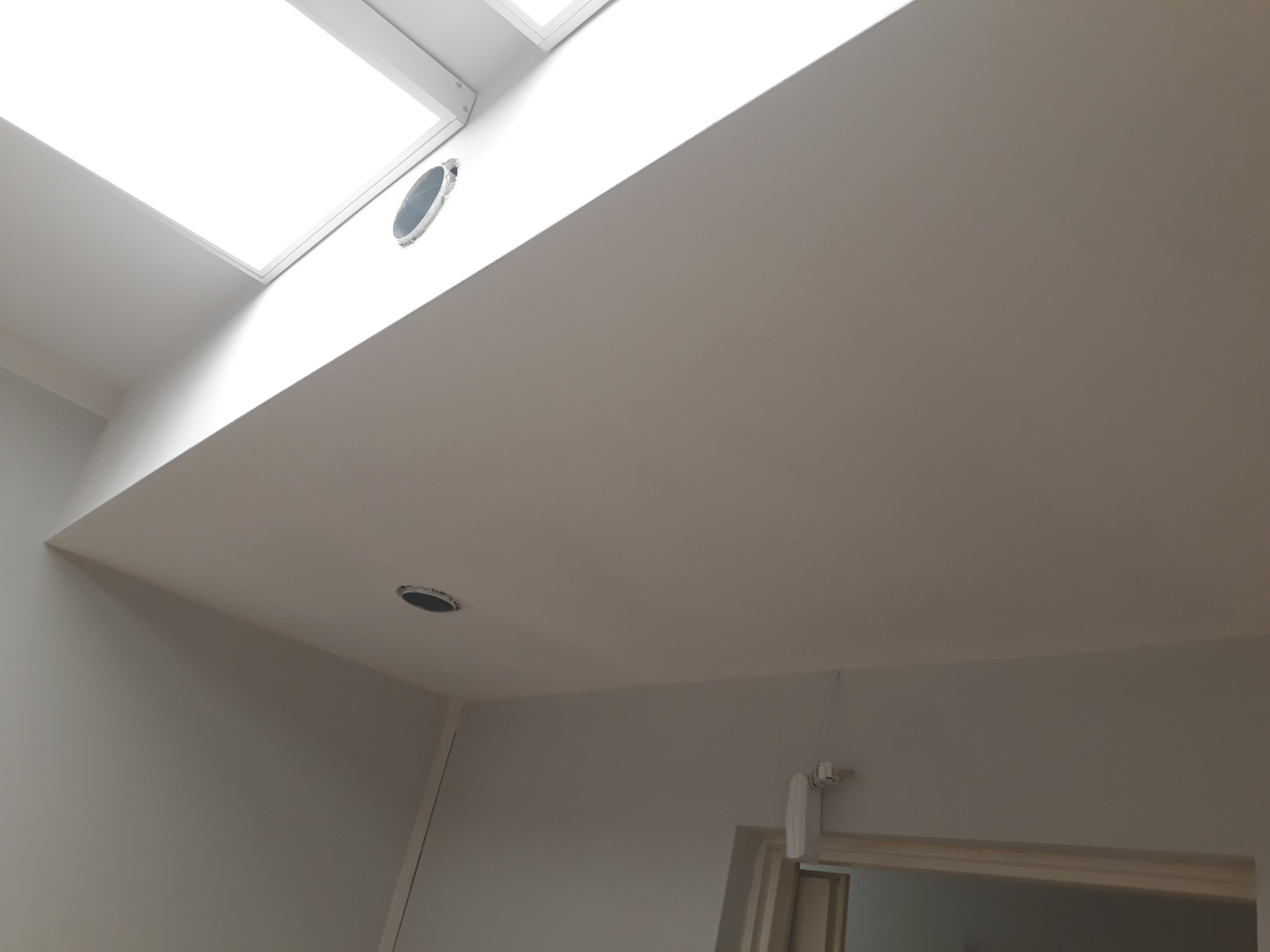 Wnętrze po remoncie, widok na gładki, pomalowany sufit z okrągłymi otworami wentylacyjnymi, górne okno dachowe i fragment ściany z drzwiami.