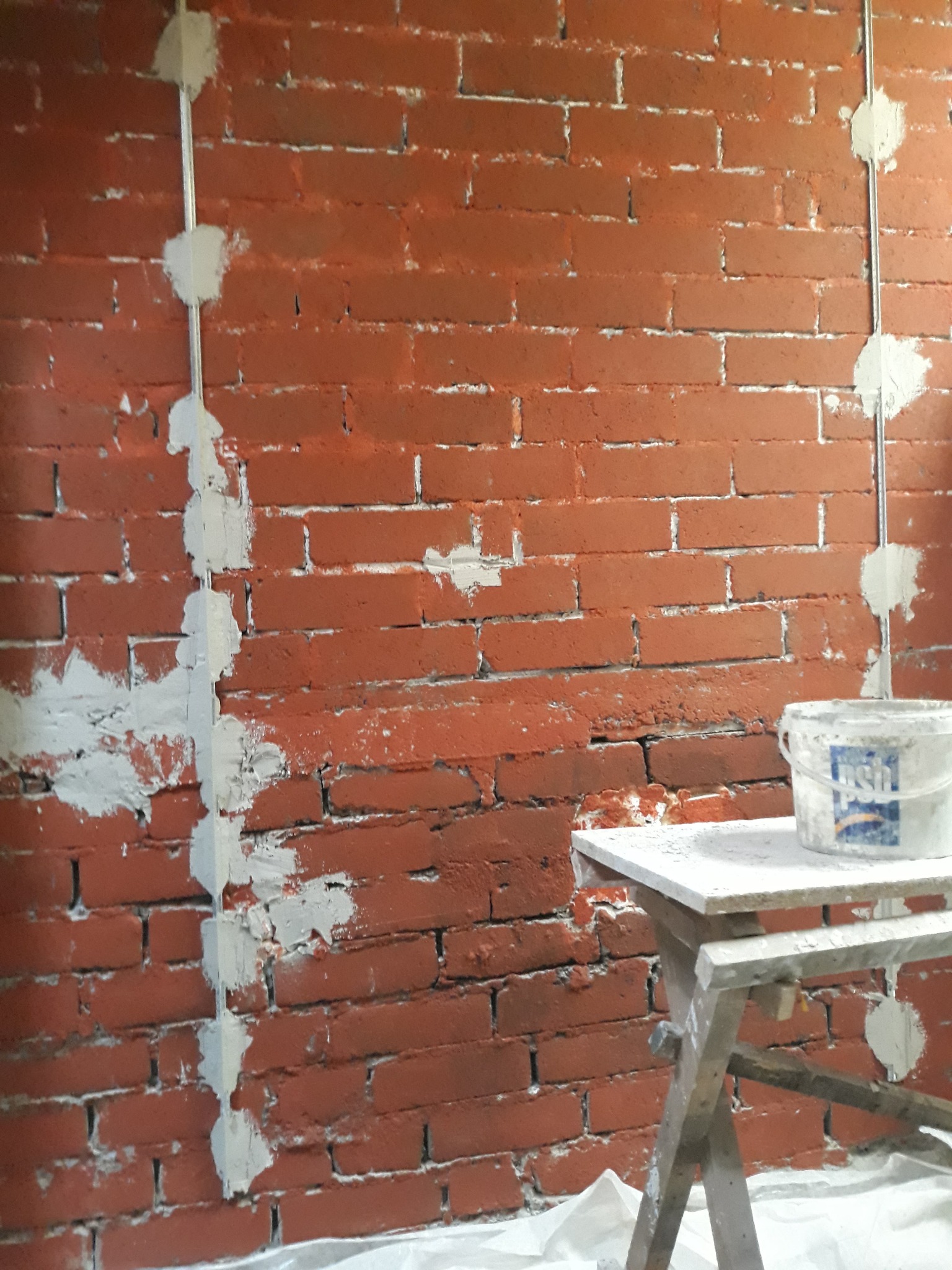 Czerwona ściana z cegły, częściowo pokryta białą zaprawą, z widocznymi przewodami elektrycznymi zabezpieczonymi gipsem i wiaderkiem farby na drewnianym taborecie.