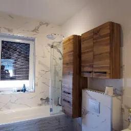 Nowoczesna łazienka z marmurowymi płytkami, drewnianymi szafkami, okrągłym lustrem i wanną z parawanem, dodatkowo dekoracje roślinne.