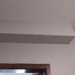 Pomalowany na biało fragment sufitu z obniżeniem, widoczny narożnik ściany i fragment ościeżnicy drzwiowej z ciemnego drewna.