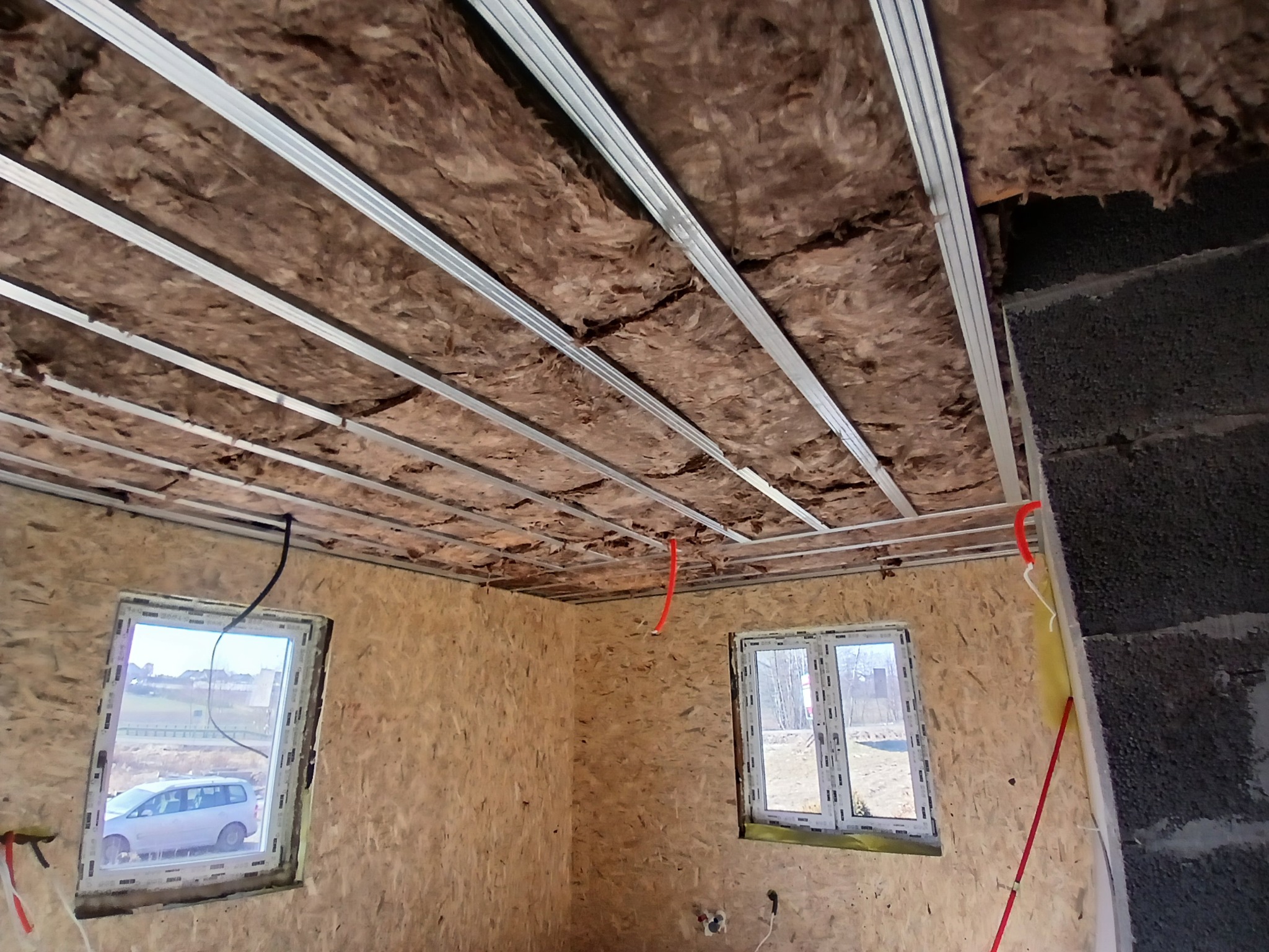 Wnętrze w trakcie prac wykończeniowych: sufit z izolacją wełną mineralną i metalowym stelażem, ściany z płyt OSB i fragment ściany z bloczków betonowych, widoczne okna.