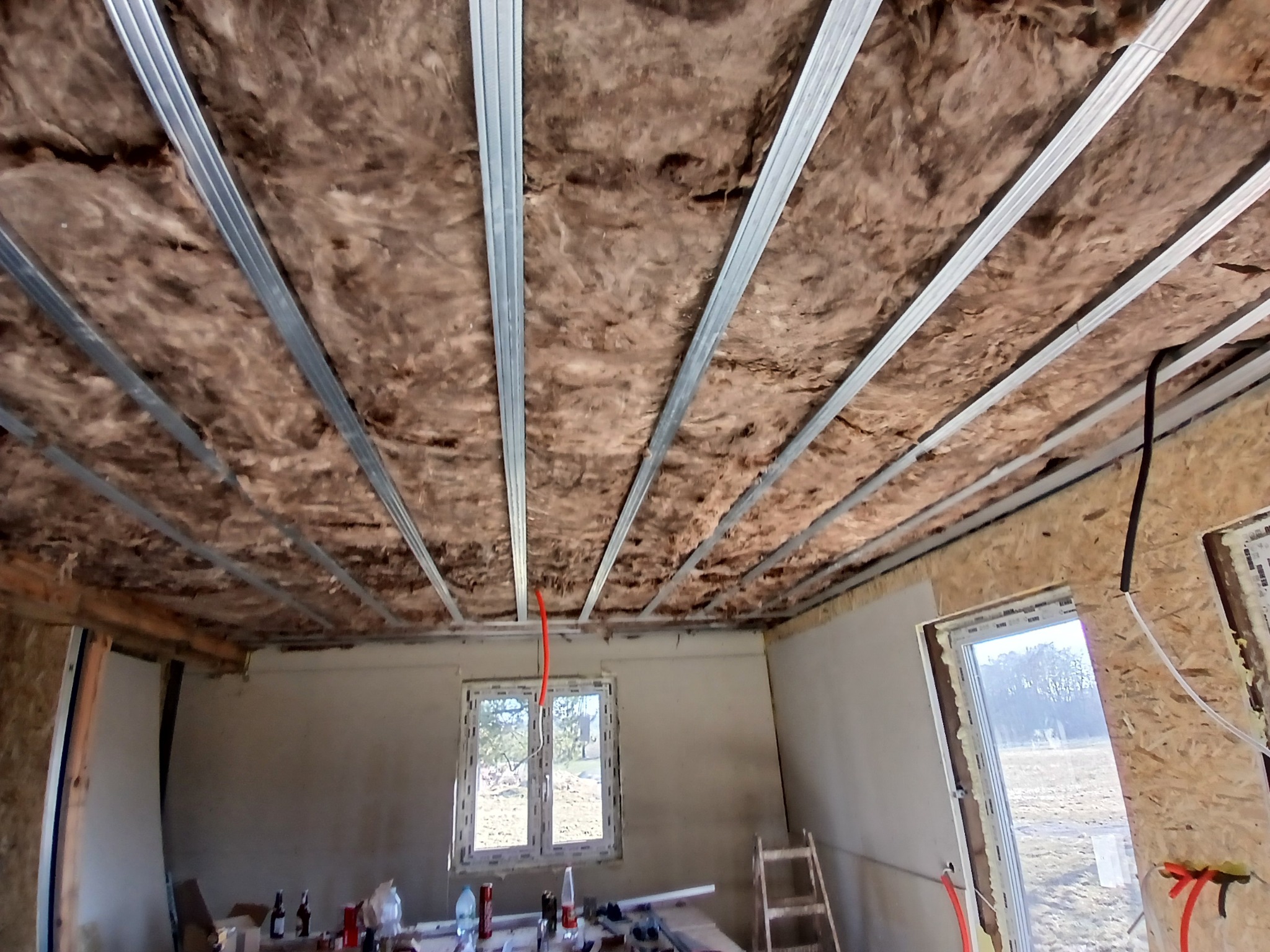 Montaż izolacji z wełny mineralnej i metalowych profili na suficie podwieszanym w surowym wnętrzu remontowanego pomieszczenia.