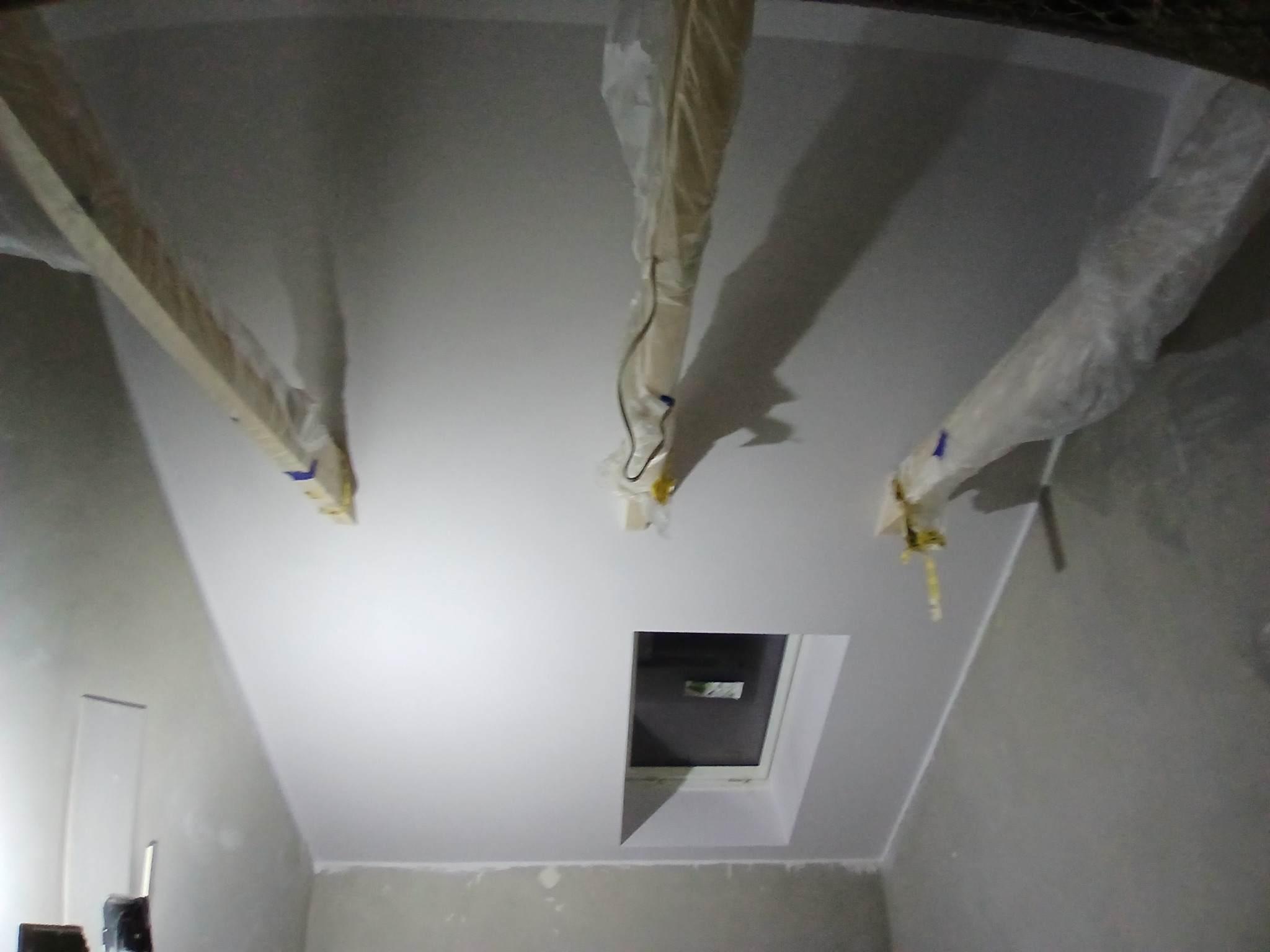 Poddasze w trakcie prac wykończeniowych: świeżo pomalowany biały sufit z oknem dachowym, zabezpieczone folią elementy konstrukcyjne, ściany przygotowane do dalszych prac.