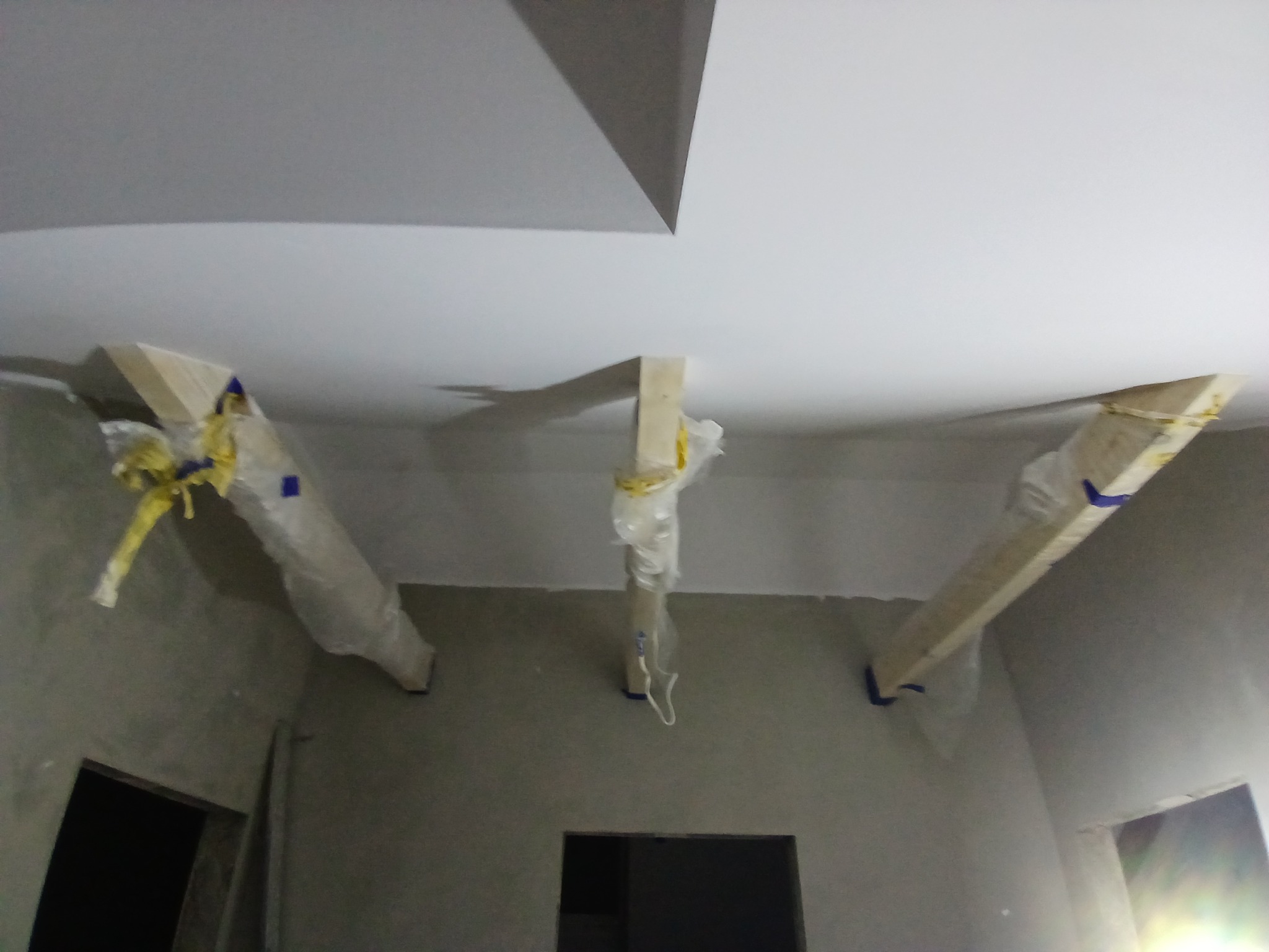 Surowy pokój z trzema drewnianymi belkami podtrzymującymi sufit, zabezpieczonymi folią i taśmą malarską, przygotowany do dalszych prac wykończeniowych.