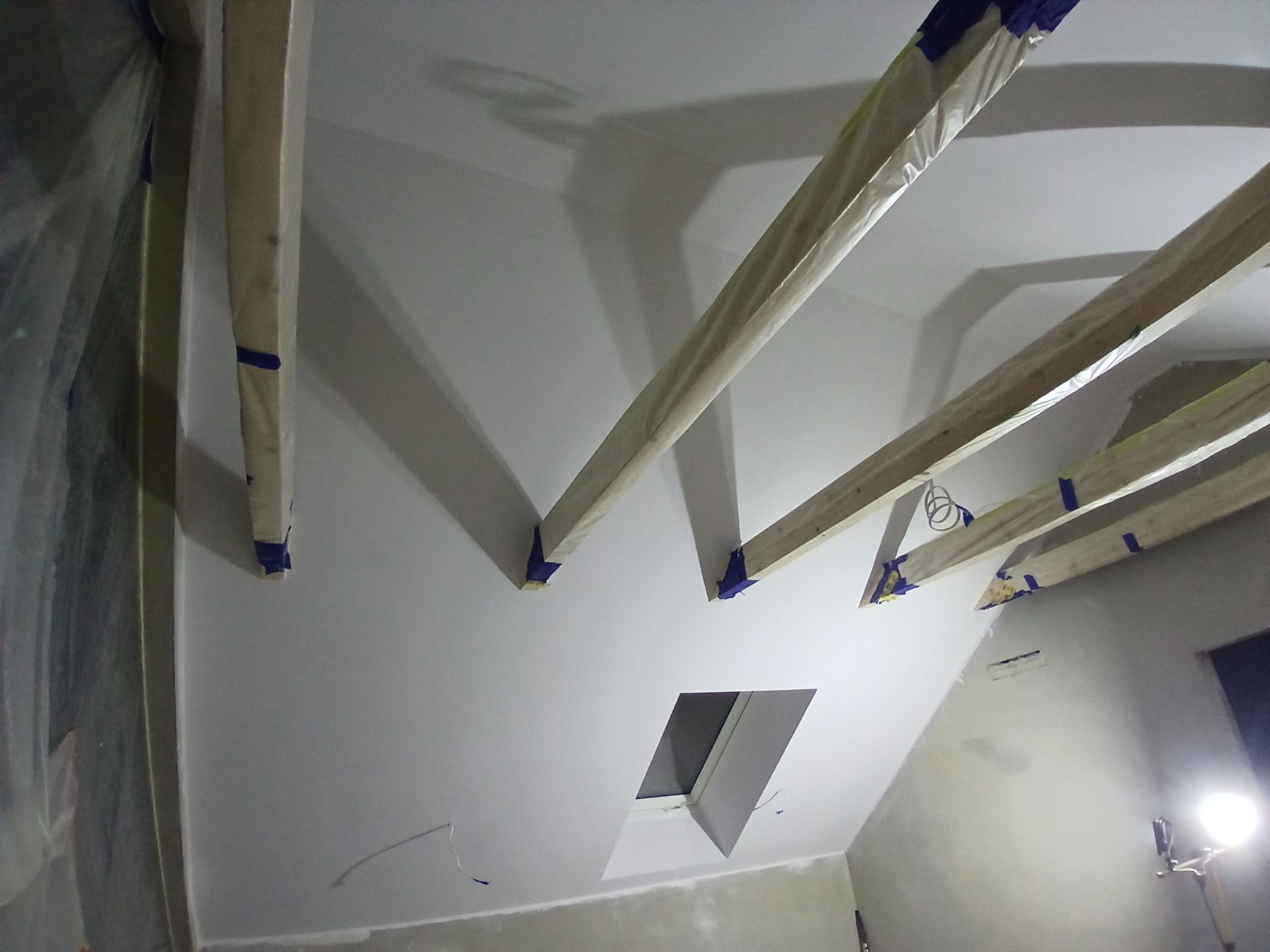 Poddasze po szpachlowaniu i malowaniu, z widocznymi drewnianymi belkami zabezpieczonymi folią i taśmą malarską, okno dachowe i ślady po kablach.