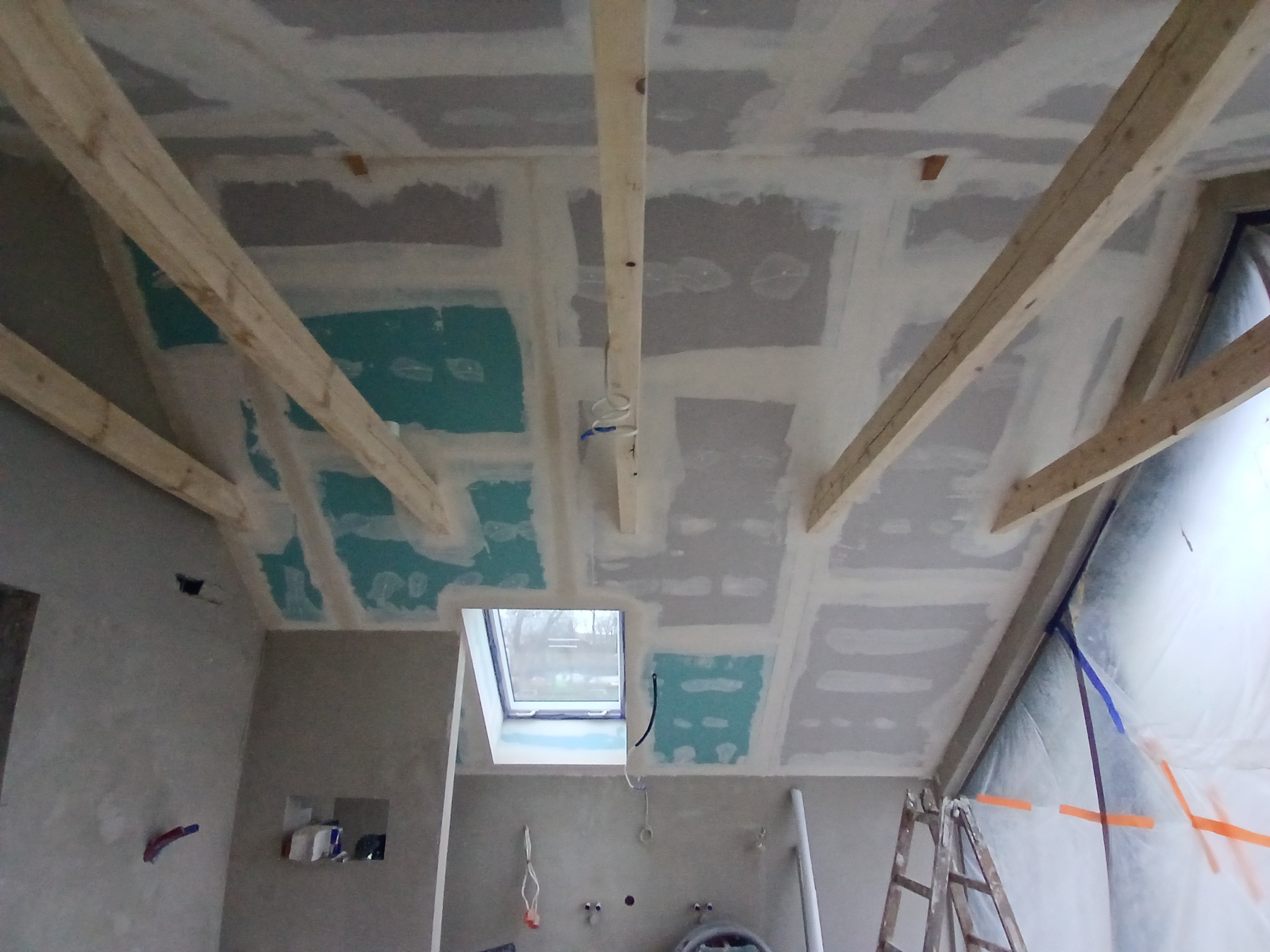 Poddasze w trakcie wykańczania: widoczne drewniane belki stropowe, płyty kartonowo-gipsowe częściowo zamontowane i zaszpachlowane, okno dachowe, ściany przygotowane do dalszych prac wykończeniowych.