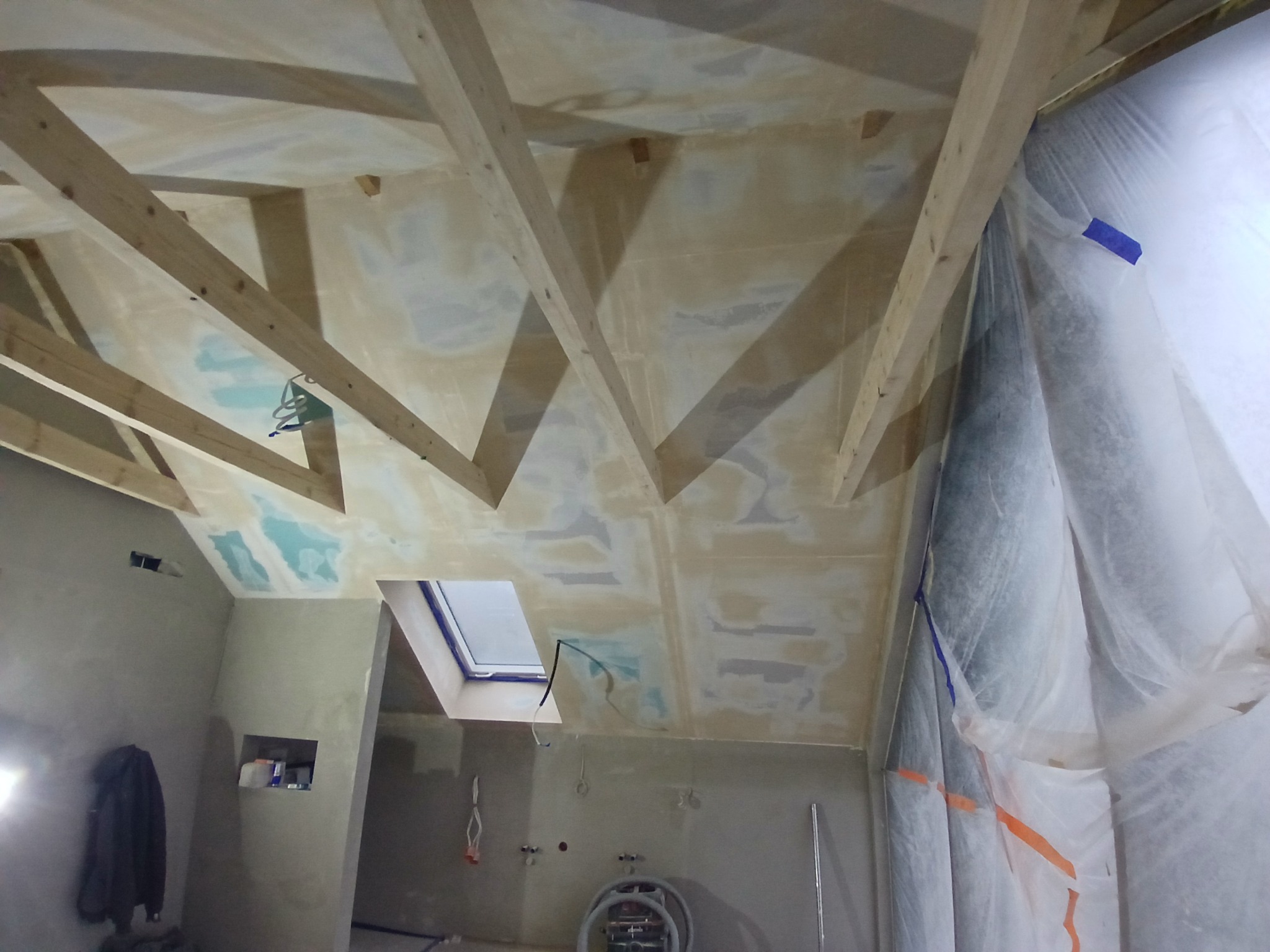 Wnętrze w trakcie prac wykończeniowych: surowy strop z drewnianą konstrukcją, częściowo zakryty folią ochronną, ściany po wstępnym wygładzeniu, widoczne okno dachowe i instalacje.