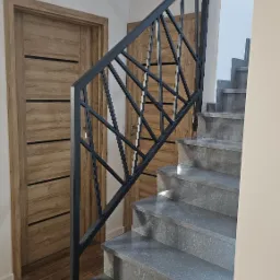 Nowoczesne schody z szarymi stopniami i czarną, geometryczną konstrukcją zabezpieczającą, widoczne drewniane drzwi w tle.