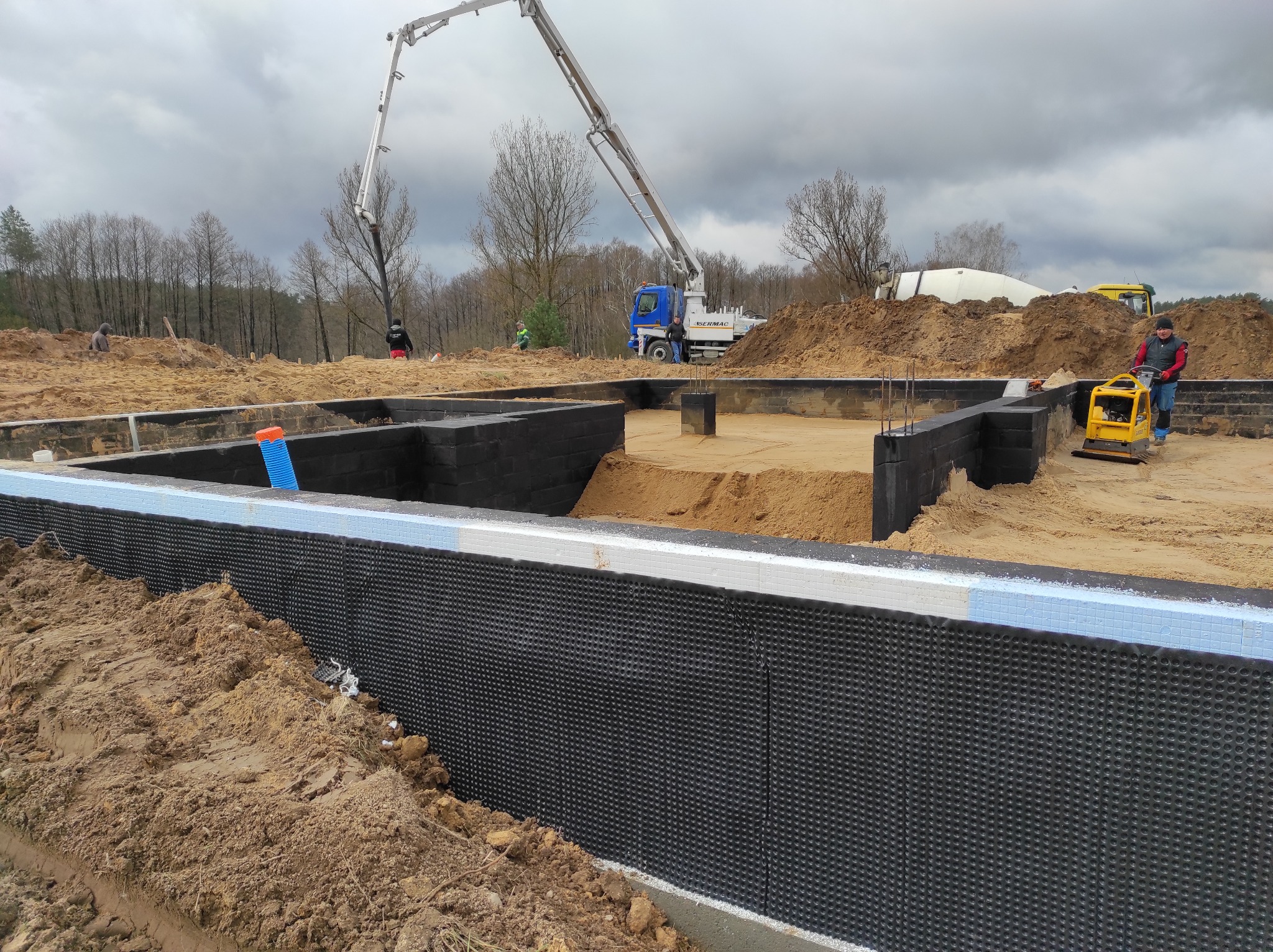 Budowa fundamentów: wylane ławy fundamentowe, izolacja pionowa ścian fundamentowych z folii kubełkowej, zagęszczarka wibracyjna, pompa do betonu i betoniarka w tle, teren budowy w trakcie prac.