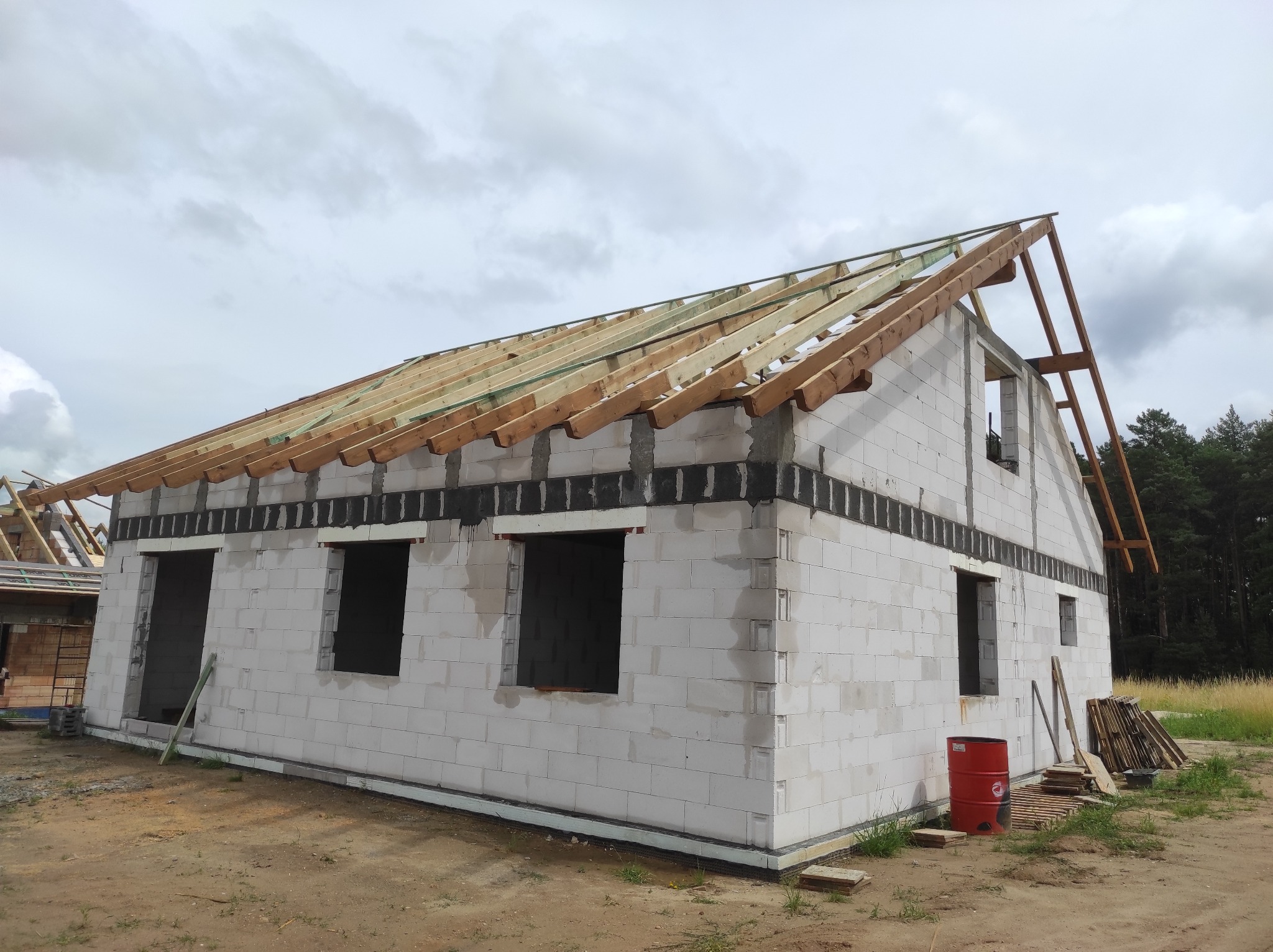 Surowy stan domu z białych bloczków, widoczne niezadaszone więźby dachowe, czerwona beczka i ułożone deski na ziemi