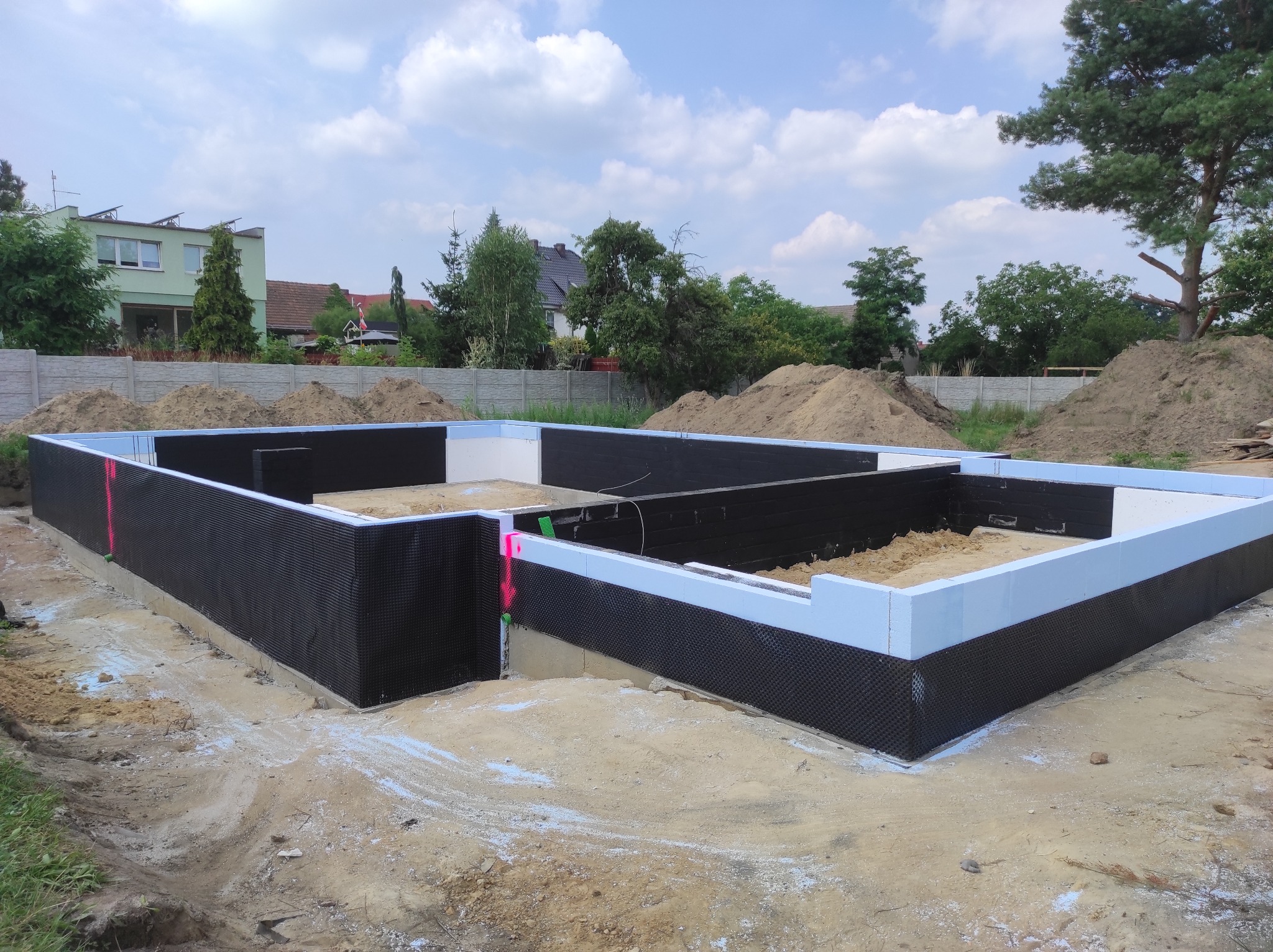 Fundament domu murowanego w trakcie budowy, zabezpieczony czarną folią kubełkową i białym styropianem, widoczne ślady wapna na ziemi, w tle hałdy ziemi i domy jednorodzinne.