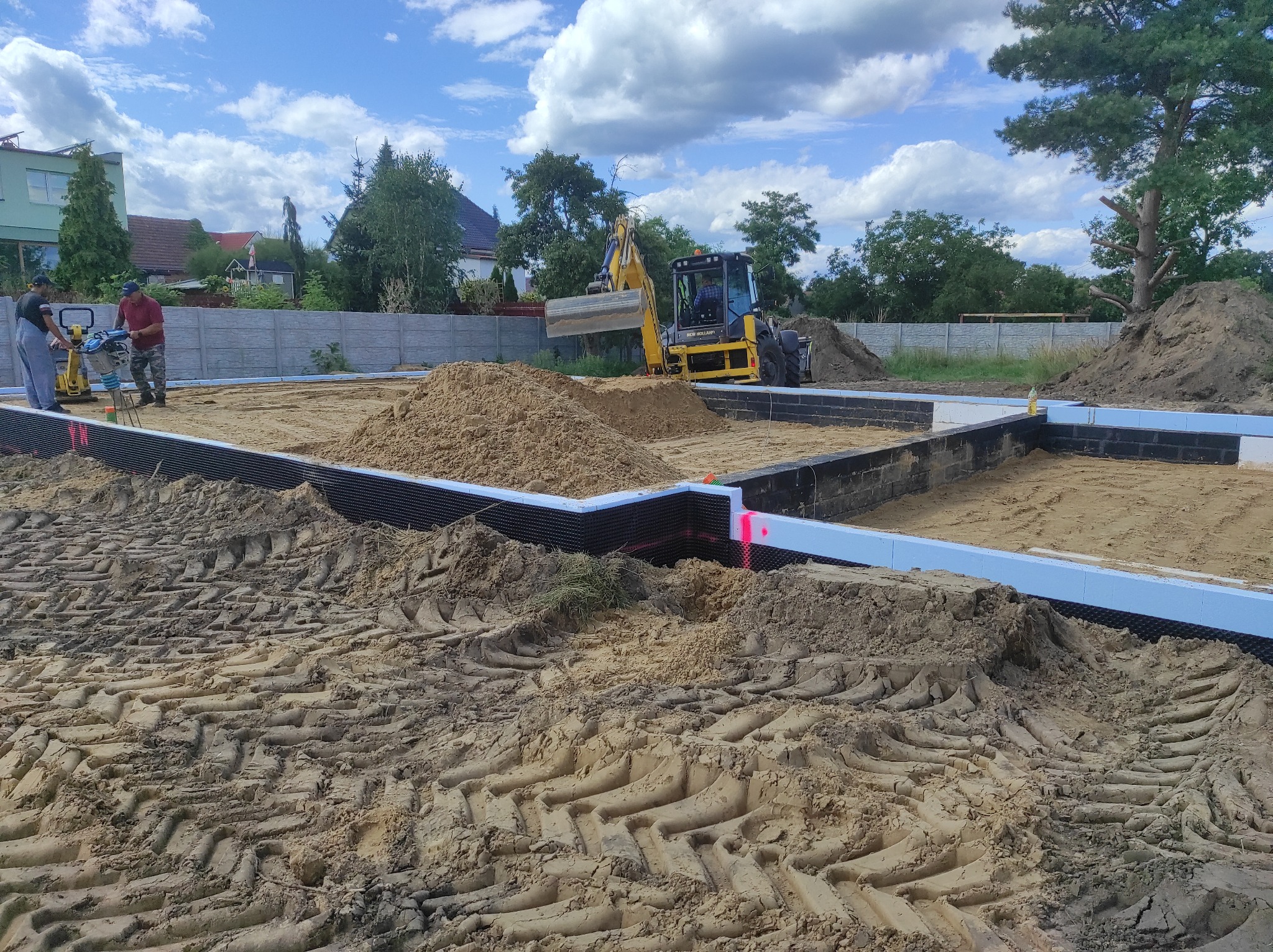 Fundamenty domu z izolacją termiczną, wykopany teren, pracownicy z niwelatorem i ubijarką, żółta koparka w tle, słoneczny dzień.