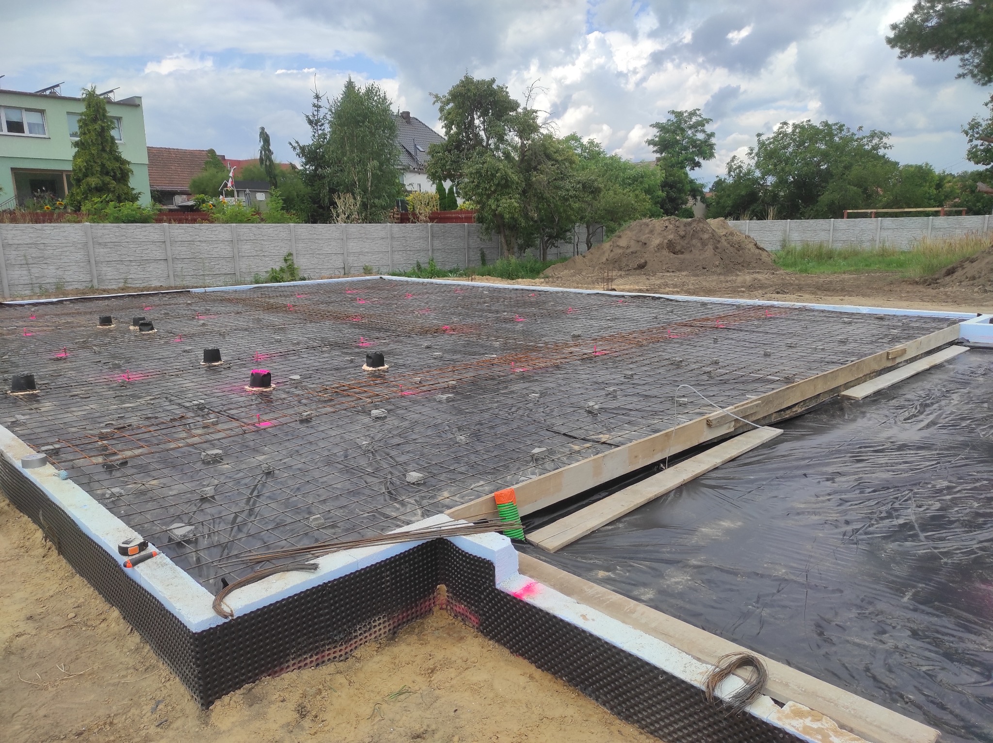 Przygotowanie fundamentów pod budowę: zbrojenie, izolacja przeciwwilgociowa, szalunki, widoczne narzędzia i materiały na placu budowy.