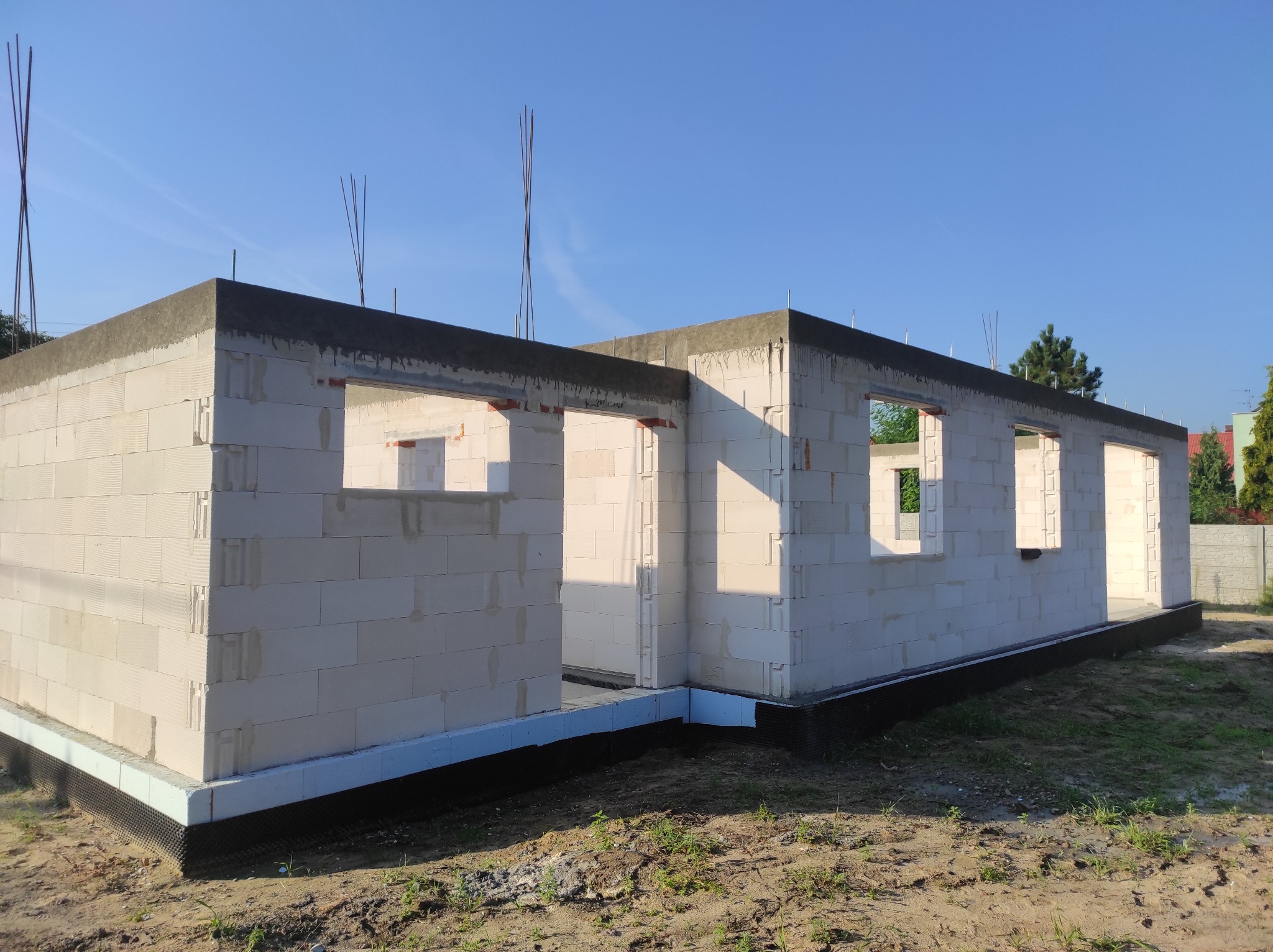 Surowy stan domu z białych bloczków, widoczne zbrojenia pionowe wystające ponad strop, fundament zabezpieczony czarną folią, w tle błękitne niebo.