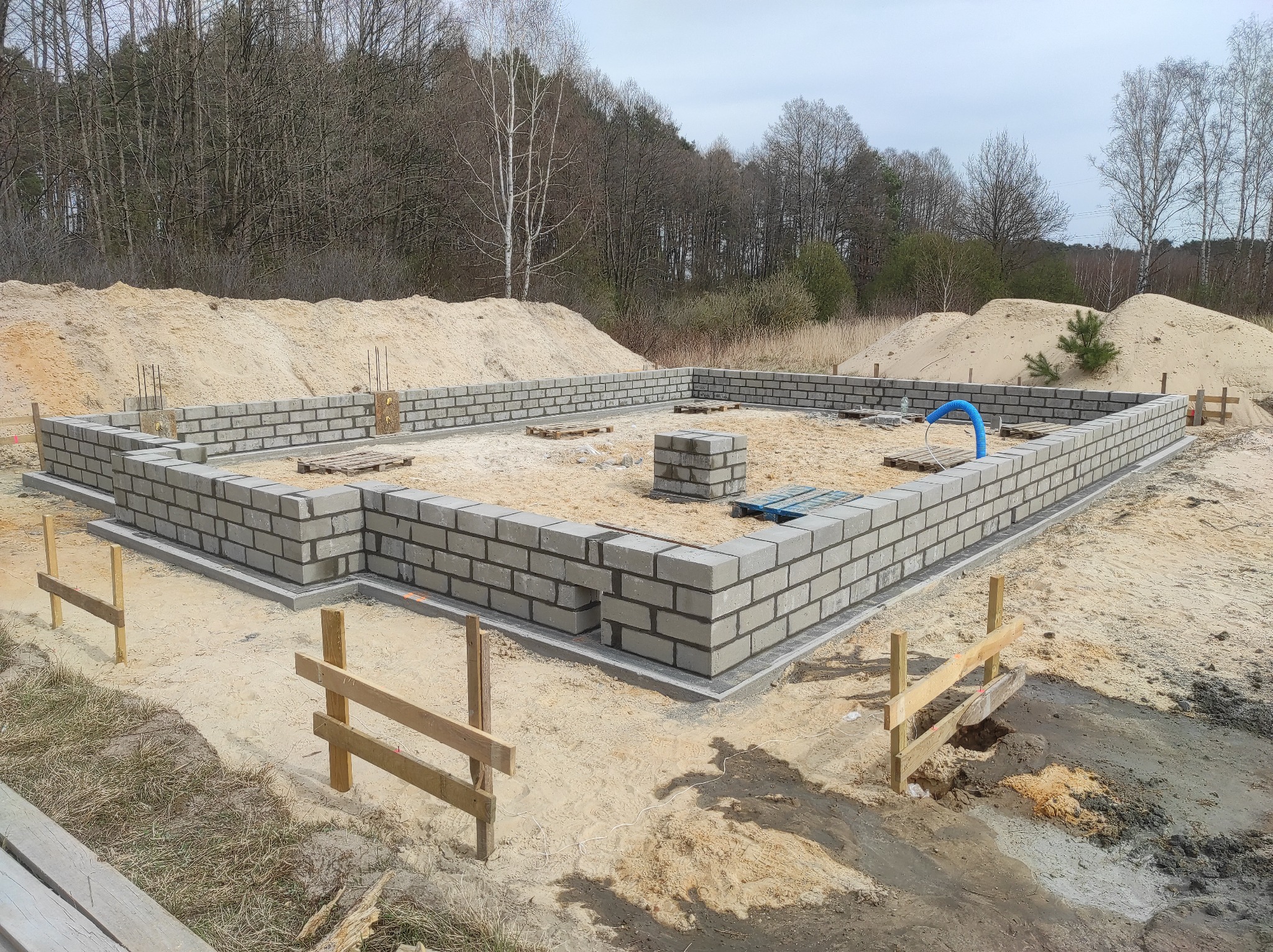Fundamenty z bloczków betonowych w trakcie budowy, widoczny niebieski wąż wentylacyjny, piasek i las w tle.