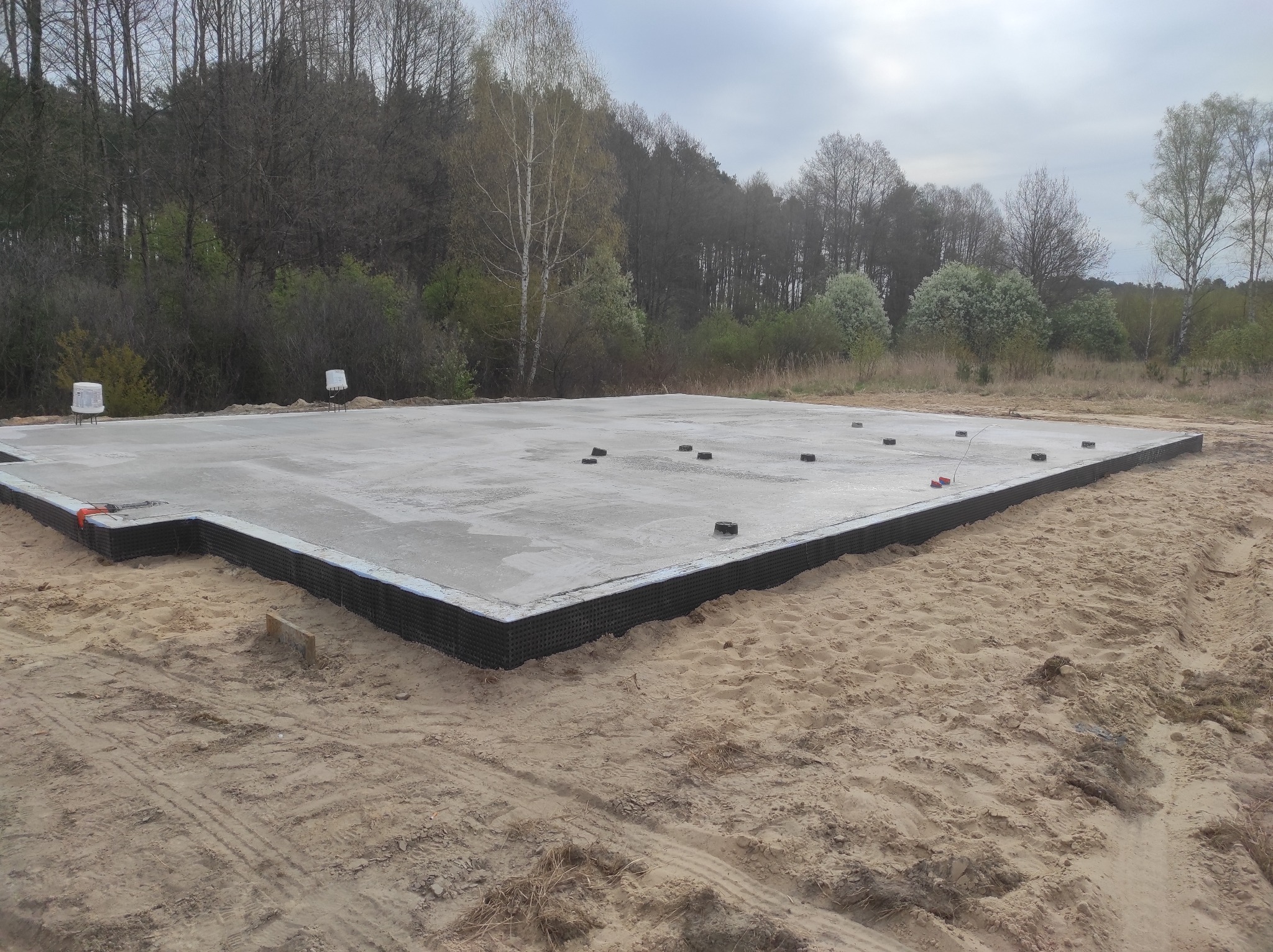 Wylana płyta fundamentowa z czarną izolacją pionową na piaszczystym terenie, widoczne czarne zaślepki na pręty zbrojeniowe, w tle drzewa bez liści i pochmurne niebo.