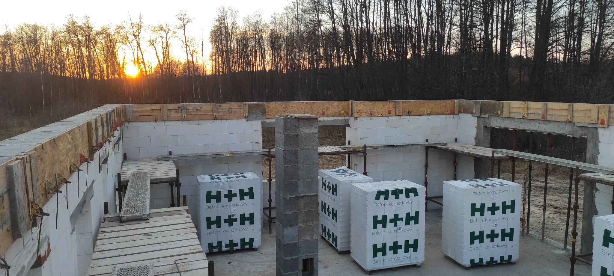 Fundamenty domu z betonu komórkowego H+H w trakcie budowy, widoczne szalunki i zbrojenia, w tle zachodzące słońce i las.