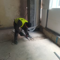 CERIGS SPÓŁKA Z OGRANICZONĄ ODPOWIEDZIALNOŚCIĄ - Pracownik w kamizelce używa młota pneumatycznego do skuwania posadzki w pomieszczeniu z instalacją elektryczną na ścianie. Widoczne gruz i surowy stan wnętrza.