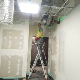 CERIGS SPÓŁKA Z OGRANICZONĄ ODPOWIEDZIALNOŚCIĄ - Pracownik na drabinie, w kamizelce odblaskowej, szpachluje ścianę w pomieszczeniu z widoczną instalacją sufitową i laserowym poziomowaniem. Etap prac wykończeniowych.