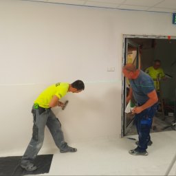 CERIGS SPÓŁKA Z OGRANICZONĄ ODPOWIEDZIALNOŚCIĄ - Dwóch mężczyzn szpachluje ścianę w jasnym pomieszczeniu, używając poziomicy laserowej. Trzeci mężczyzna w tle, drzwi z widocznym profilem aluminiowym.