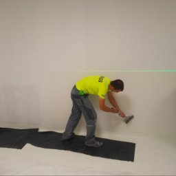 CERIGS SPÓŁKA Z OGRANICZONĄ ODPOWIEDZIALNOŚCIĄ - Pracownik w neonowej koszulce szpachluje ścianę, używając poziomicy laserowej. Na podłodze czarna folia ochronna. Widoczny fragment drugiego pracownika.