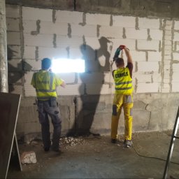 CERIGS SPÓŁKA Z OGRANICZONĄ ODPOWIEDZIALNOŚCIĄ - Dwóch pracowników w kamizelkach wierci otwór w ścianie z bloczków betonowych. Widoczne narzędzia i drabina. Praca w toku, surowy stan.