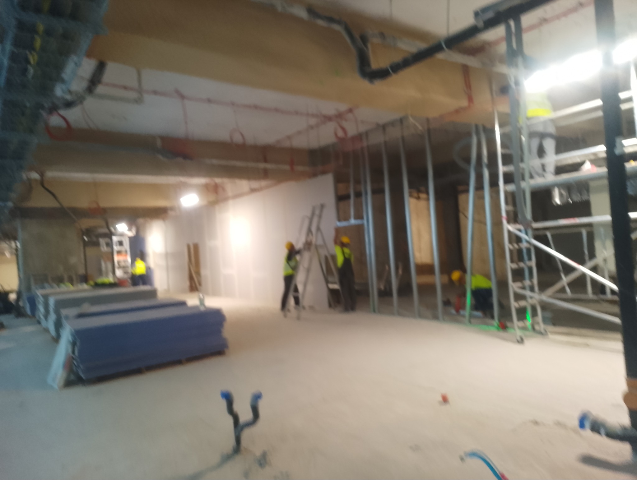 Montaż ścian działowych z płyt gipsowo-kartonowych w przestrzeni biurowej. Widoczne profile, płyty i pracownicy w odblaskowych kamizelkach. Prace w toku, surowy stan.