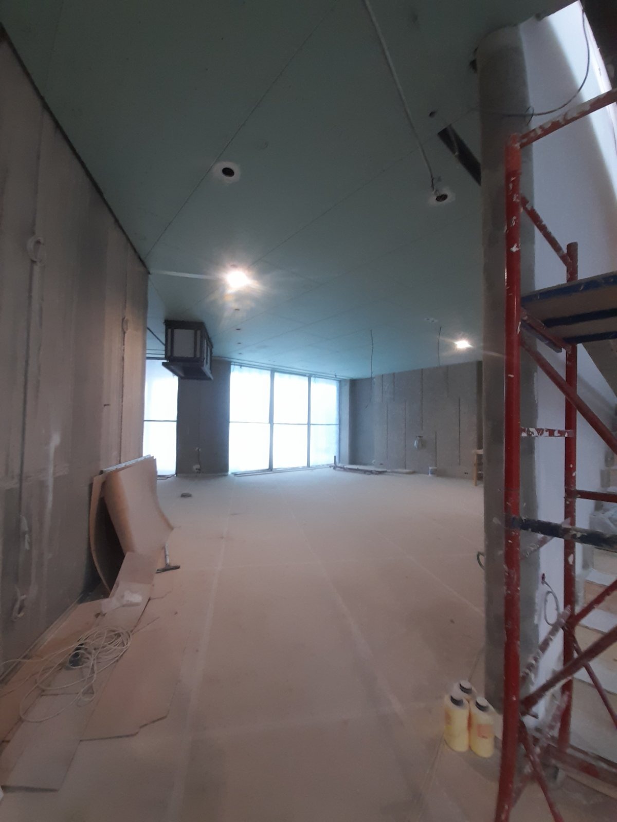 Wnętrze w trakcie prac wykończeniowych: ściany i sufit pokryte płytami gipsowo-kartonowymi, widoczne rusztowanie i materiały budowlane. Duże okna w tle.