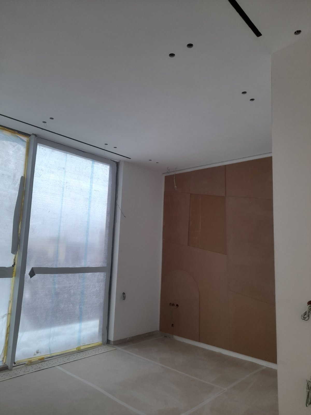 Wnętrze w trakcie prac wykończeniowych: ściana z płyt, zabezpieczone okno folią, sufit z punktowym oświetleniem. Widoczne przygotowania do dalszych prac.