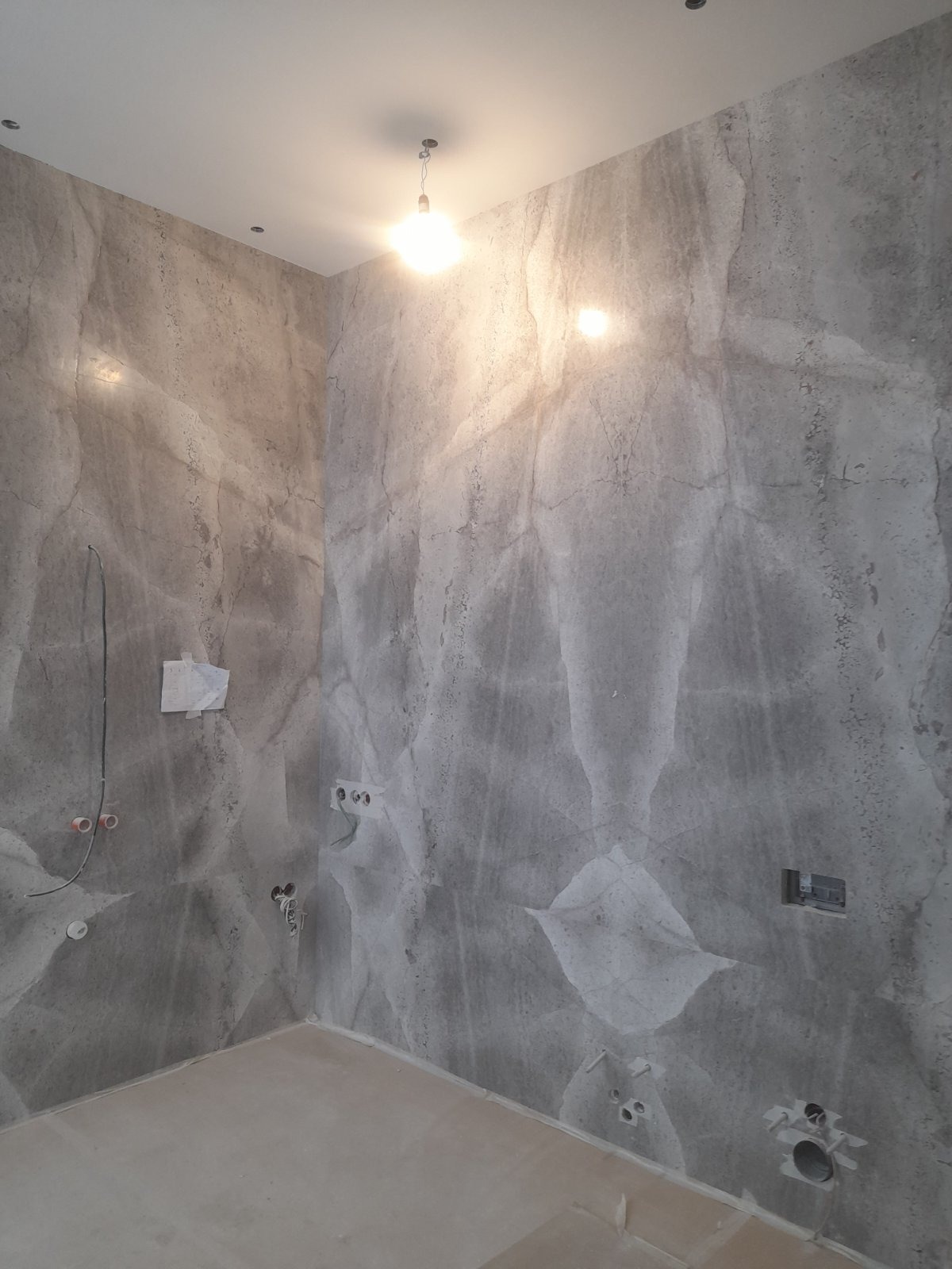 Wykończona łazienka z szarą, marmurkową okładziną ścian. Widoczne punkty instalacji elektrycznej i wodnej. Minimalistyczna lampa sufitowa.