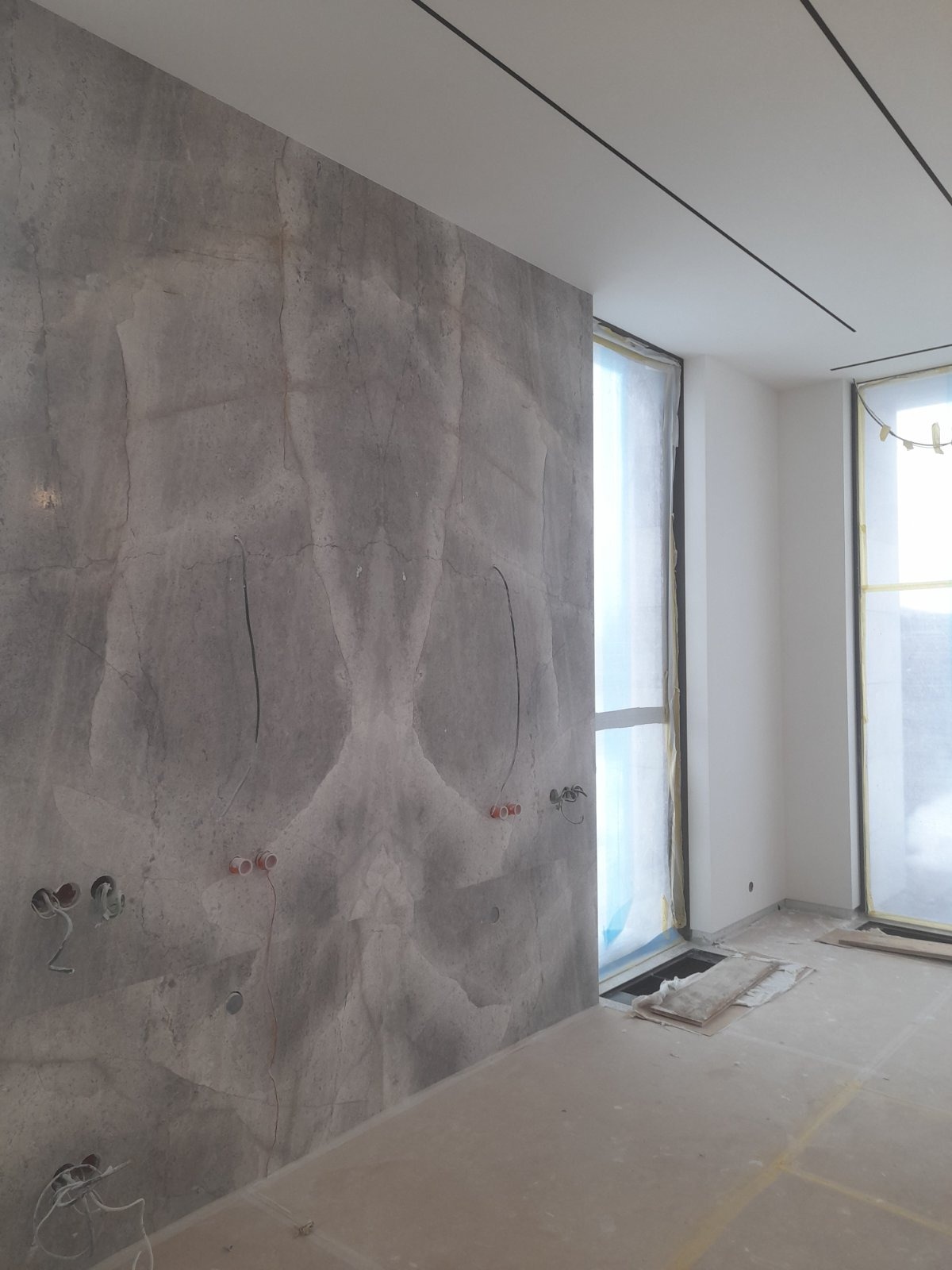 Ściana z efektem kamienia i przygotowane punkty elektryczne w jasnym wnętrzu. Okna zabezpieczone folią malarską. Minimalistyczne oświetlenie sufitowe.