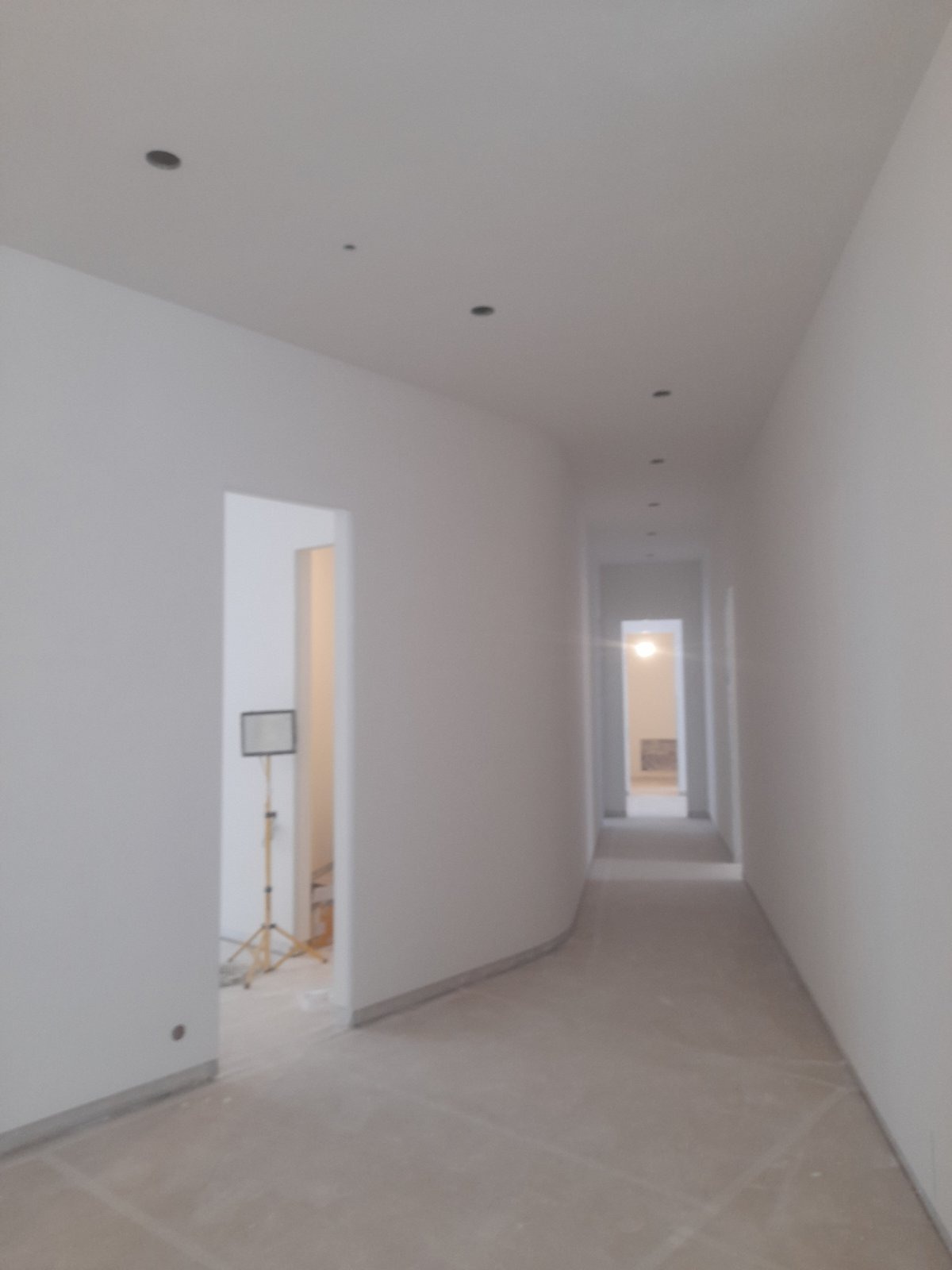 Biały korytarz po szpachlowaniu i malowaniu, widoczne otwory na oświetlenie punktowe, w tle lampa budowlana. Minimalistyczne wykończenie wnętrza.